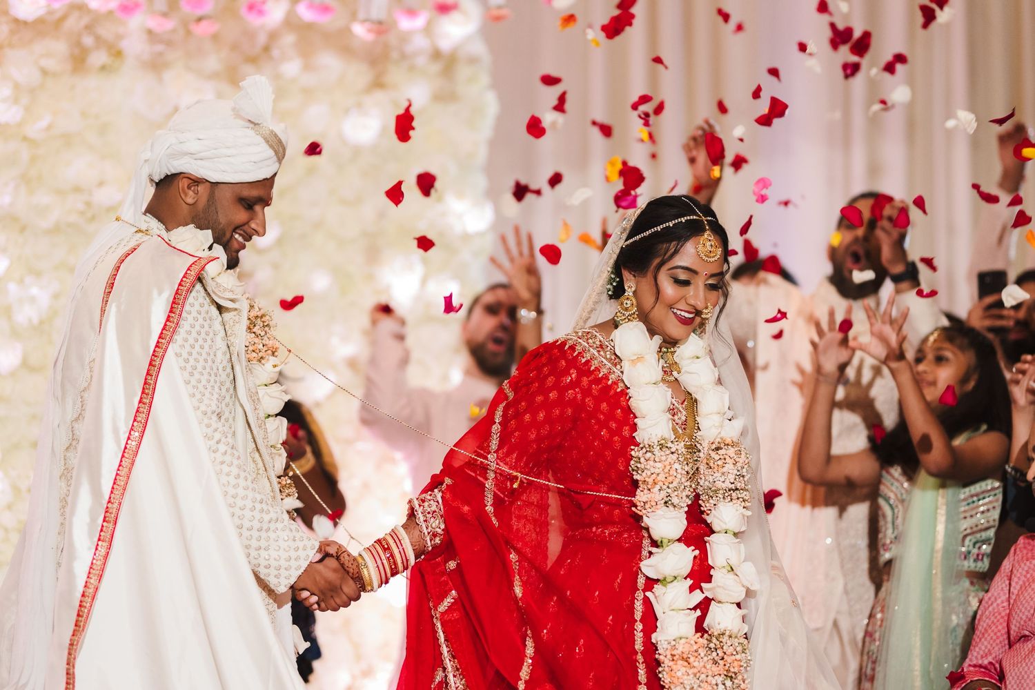 Red and white wedding celebration with rose petals falling during traditional Indian ceremony.