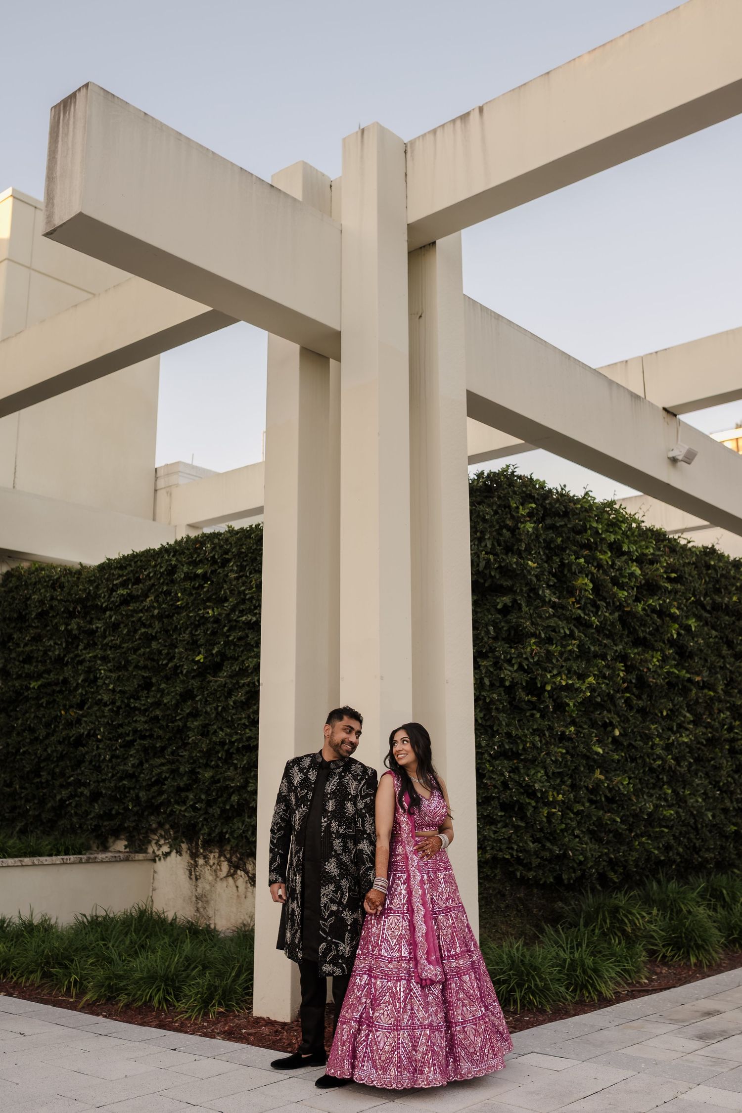A couple in traditional Indian wedding attire poses against white architectural columns and green hedges.