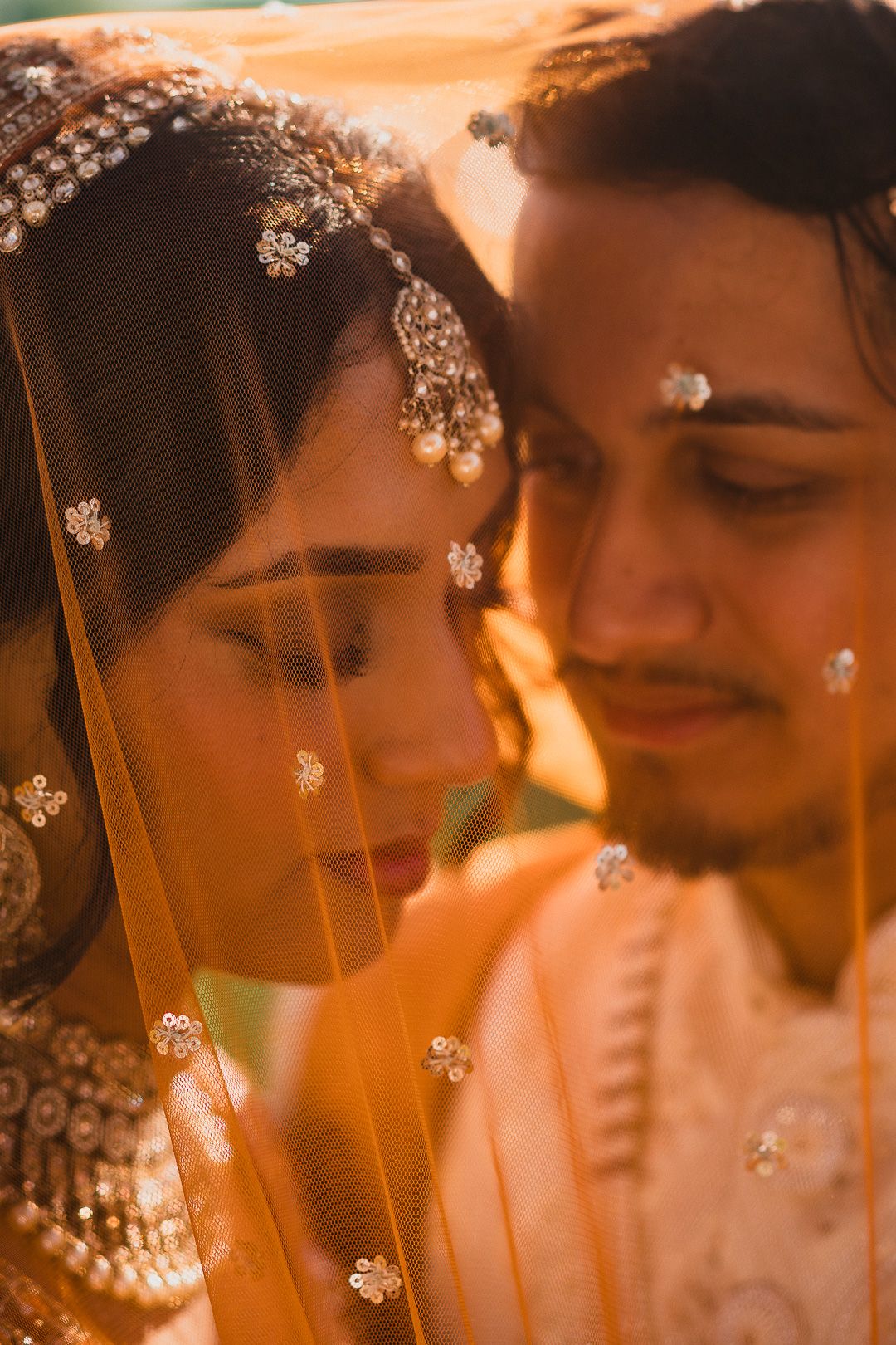 Intimate wedding portrait with traditional Indian bridal details and golden lighting through sheer orange veil.
