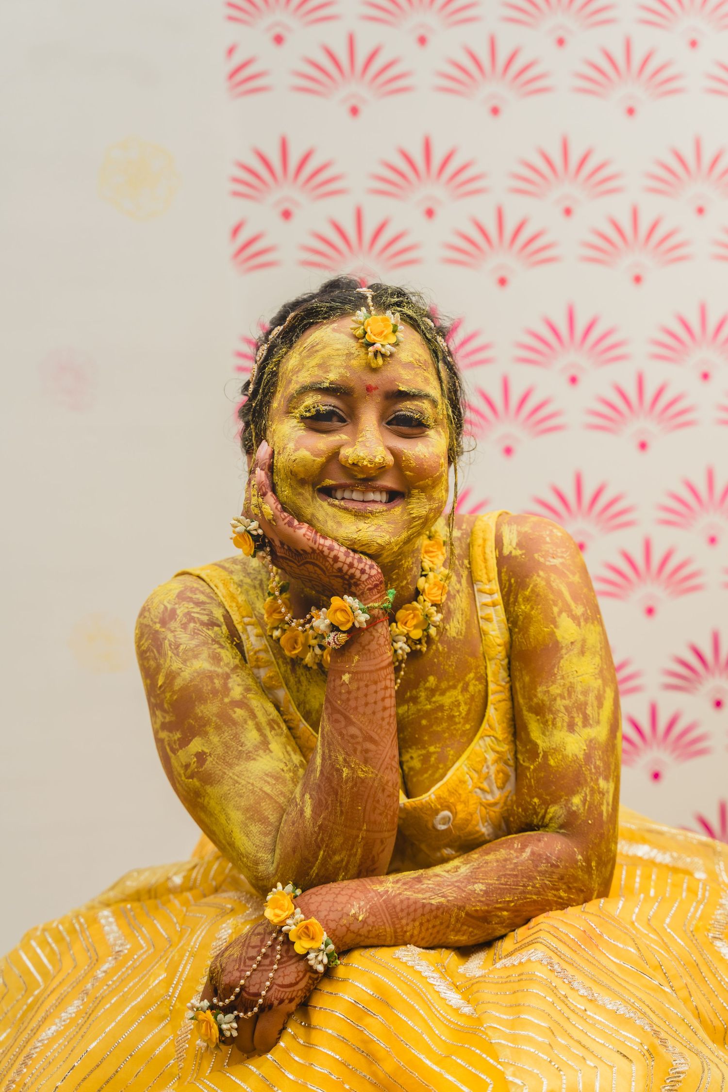 Traditional Indian bridal attire with turmeric-covered hands and floral jewelry against a pink patterned backdrop.