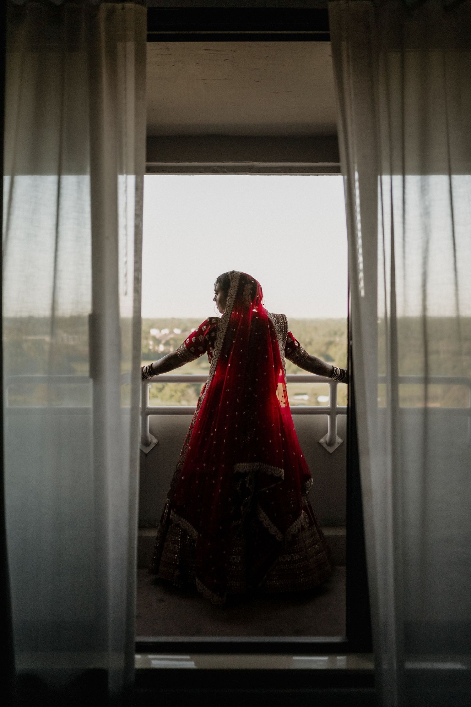 Silhouette through sheer curtains shows figure in red bridal lehenga standing on balcony.