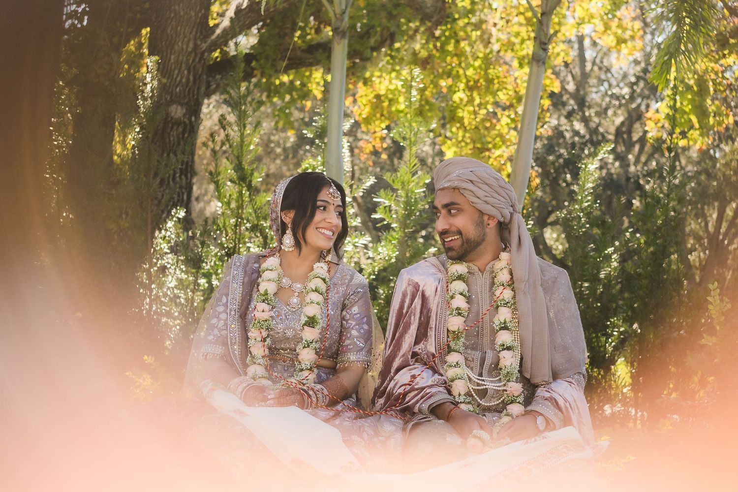 A couple in traditional Indian wedding attire sits together on a sunny day in a forest setting.