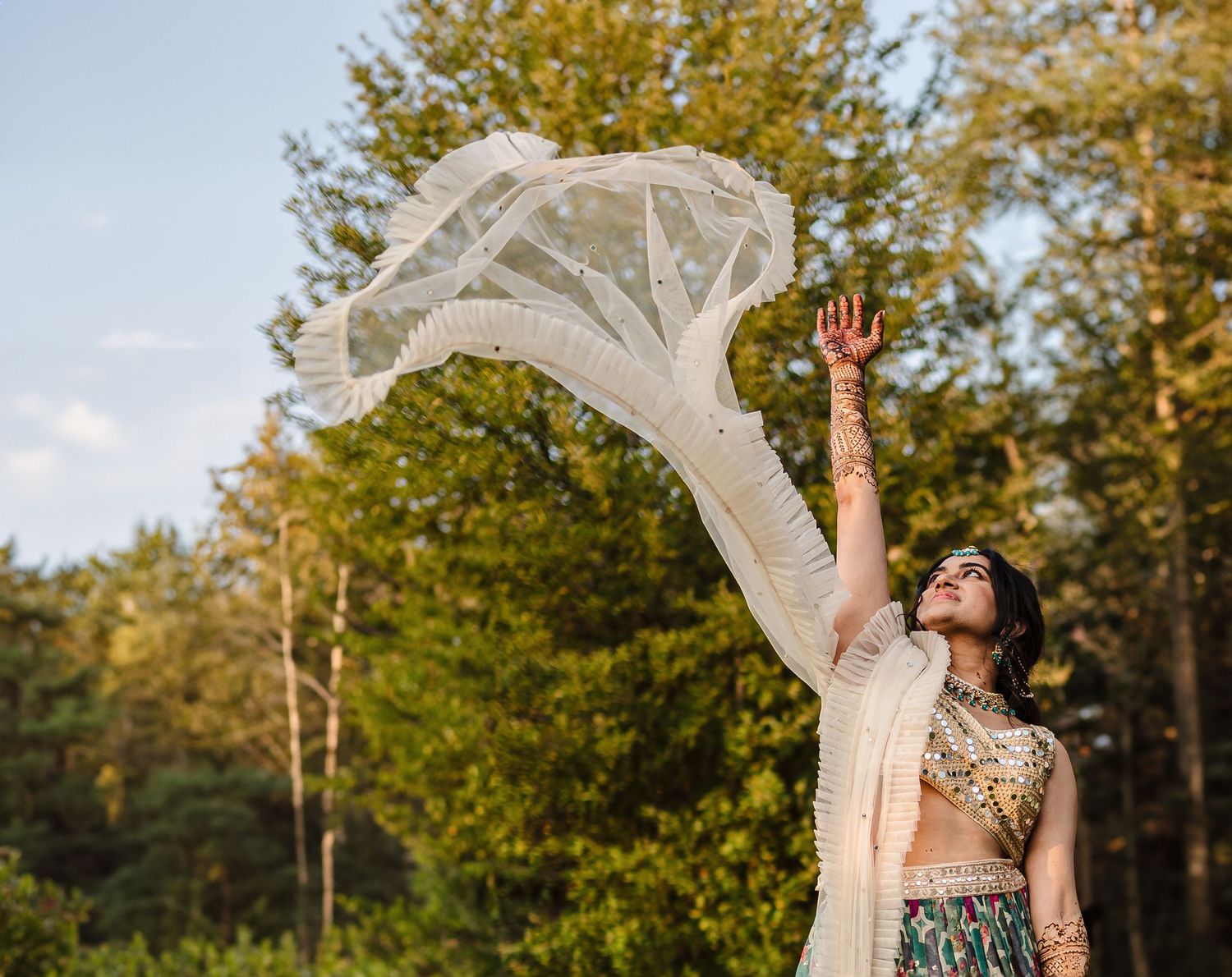 Person in fusion attire twirling white veil against autumn trees.