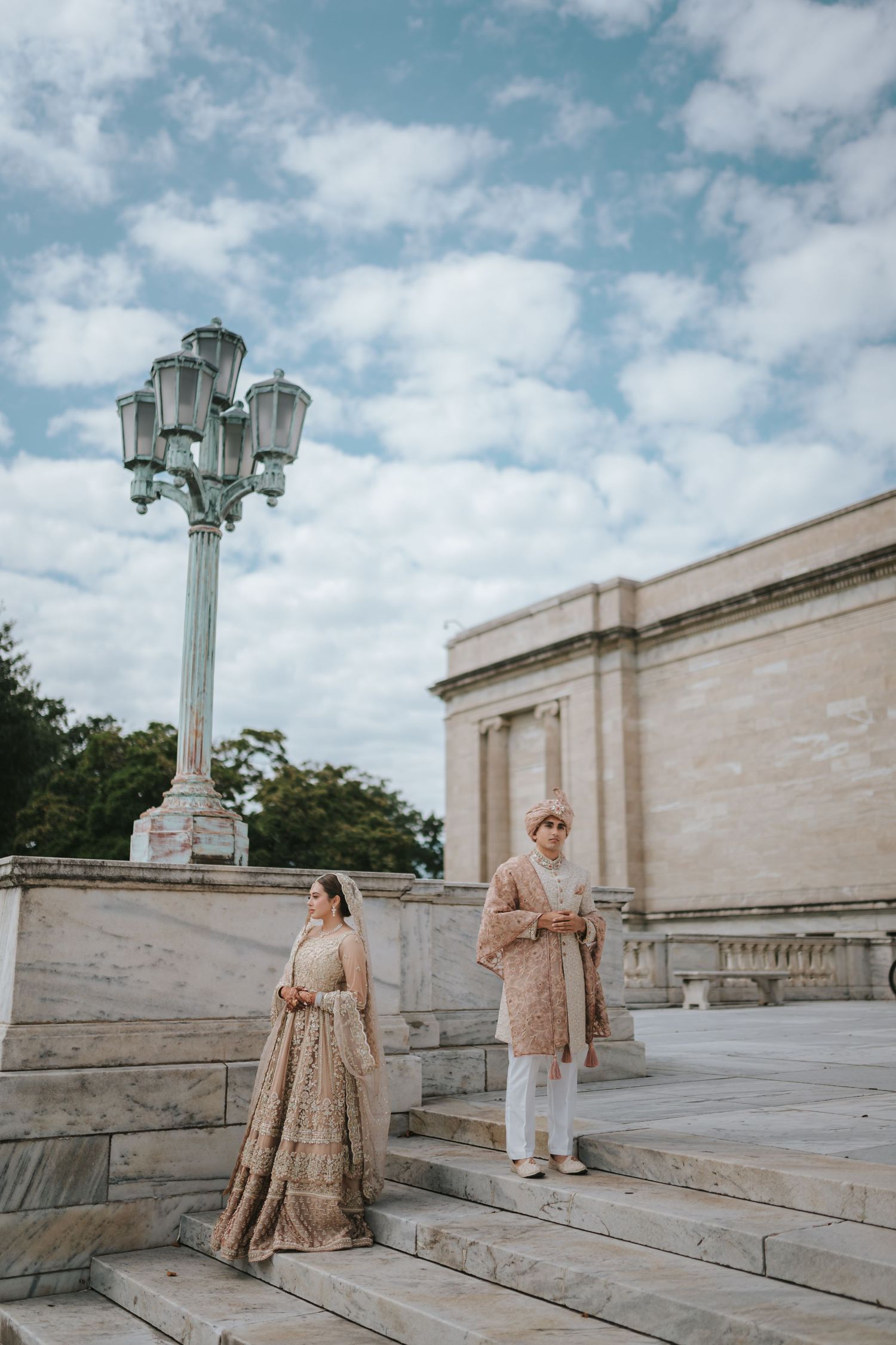 Couple in traditional South Asian wedding attire pose on stone steps near vintage lamp post.