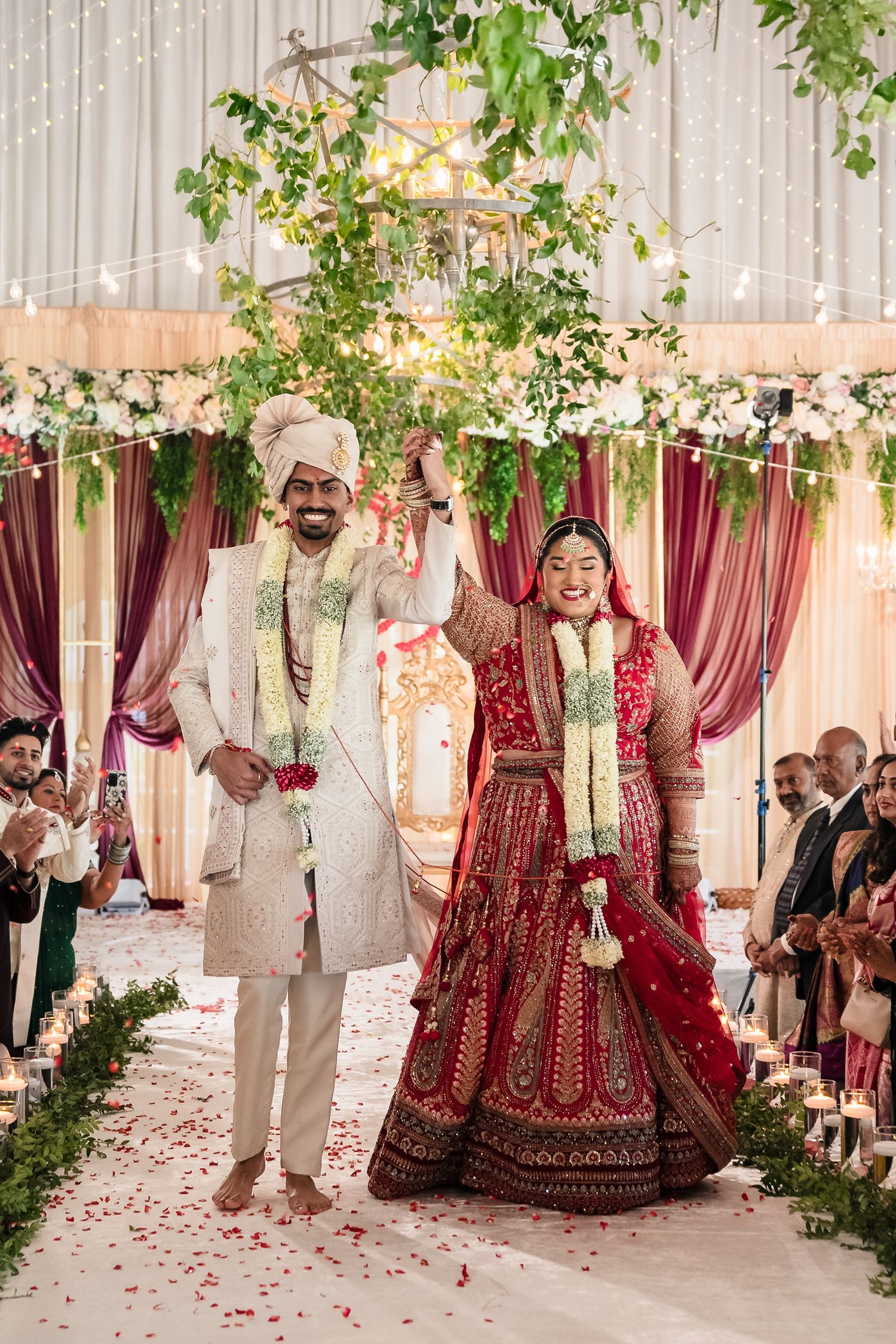 A traditional Indian wedding ceremony with couple in ornate attire standing beneath floral decor and draped burgundy fabric.