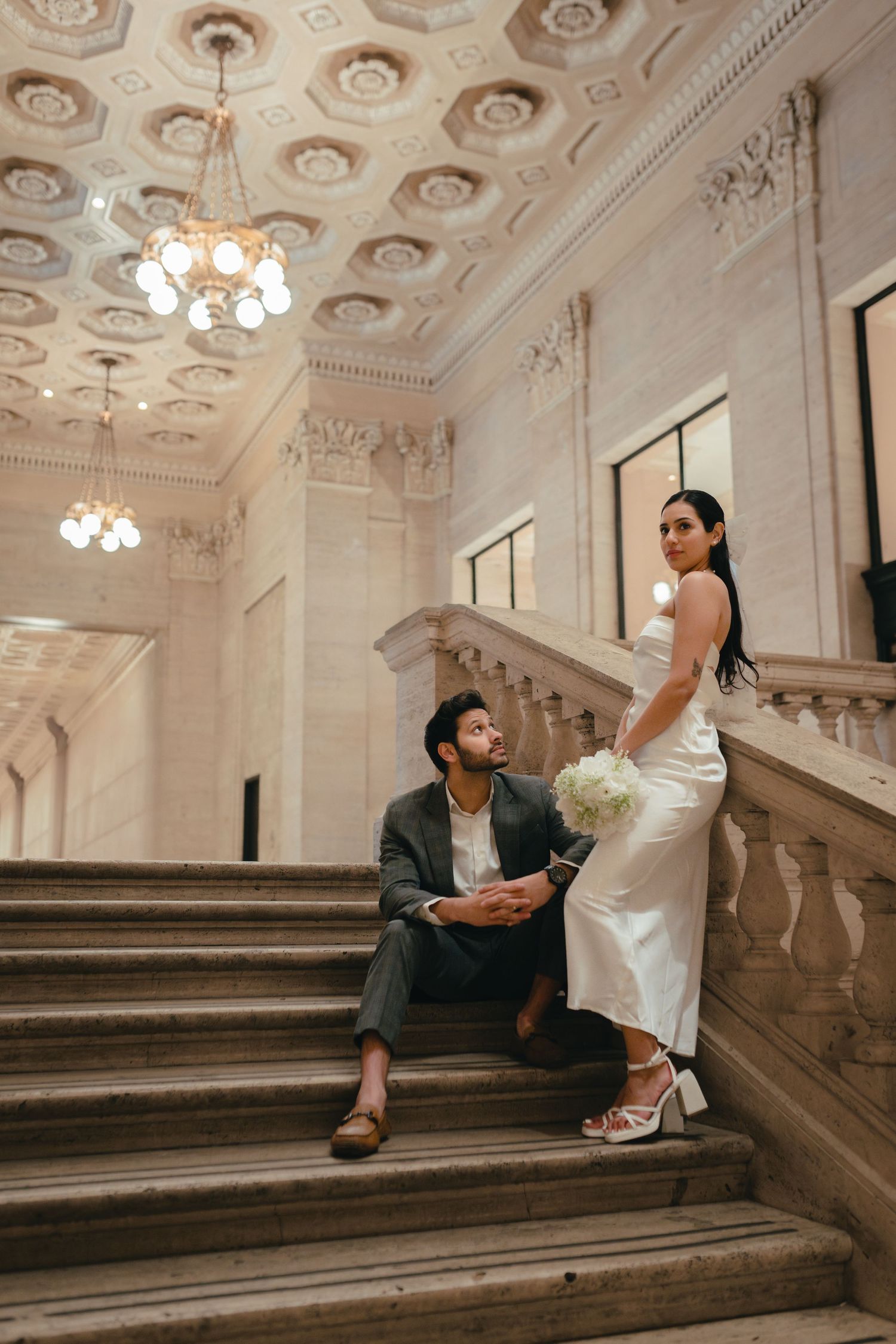 A couple poses on an ornate marble staircase in a grand building with decorative ceiling and chandeliers.
