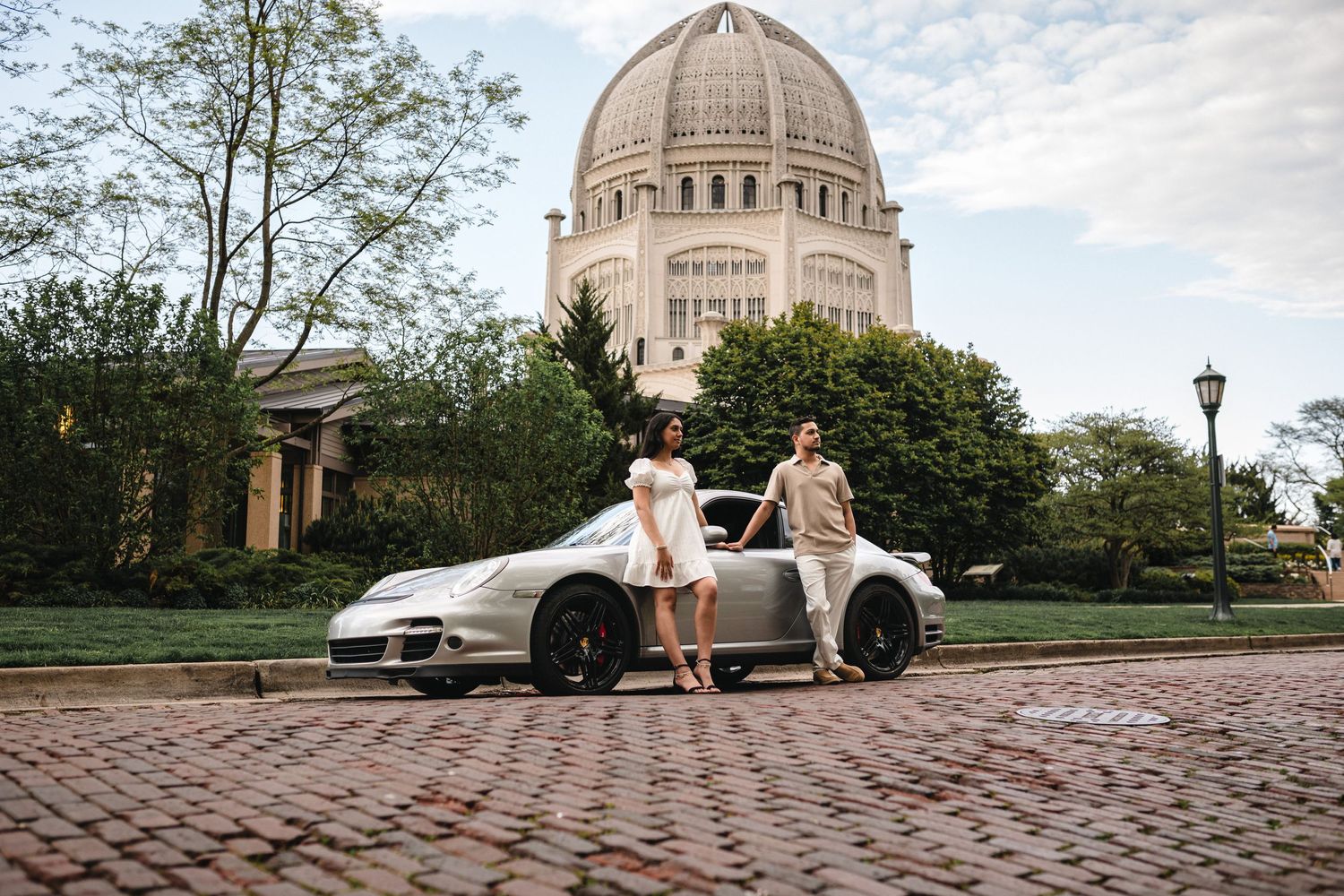 A couple sits on a vintage sports car in front of the domed Baha'i Temple on a cobblestone street.