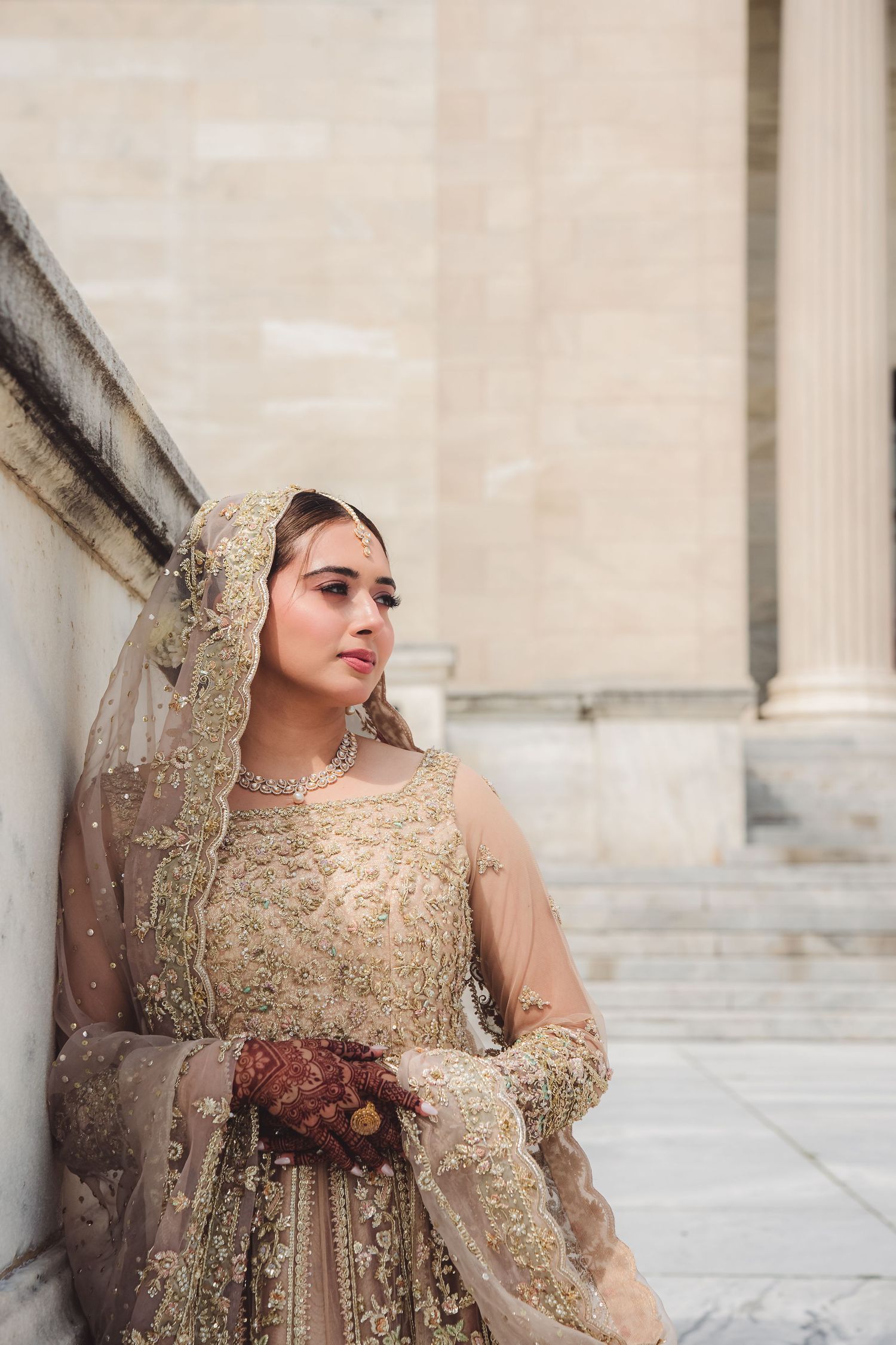 A bride in an ornate gold embroidered traditional South Asian wedding dress poses against a stone wall.