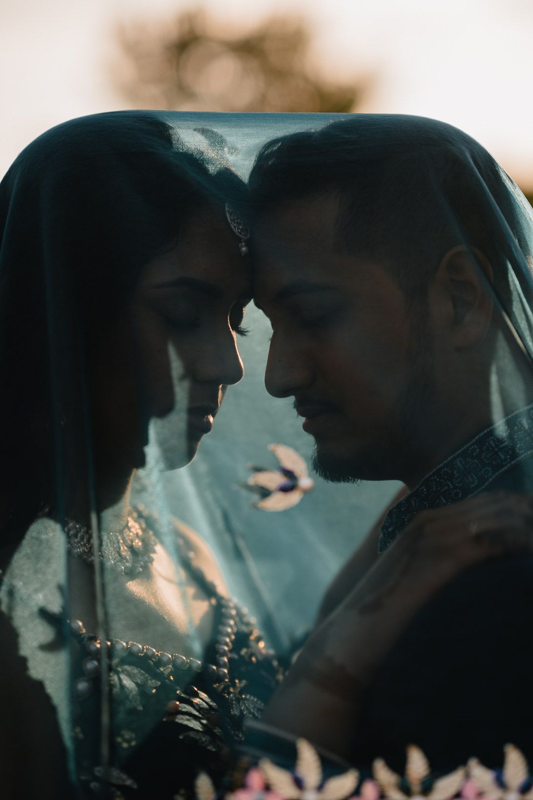A romantic silhouette of a couple in traditional attire touching foreheads through glass with a dreamy teal background.