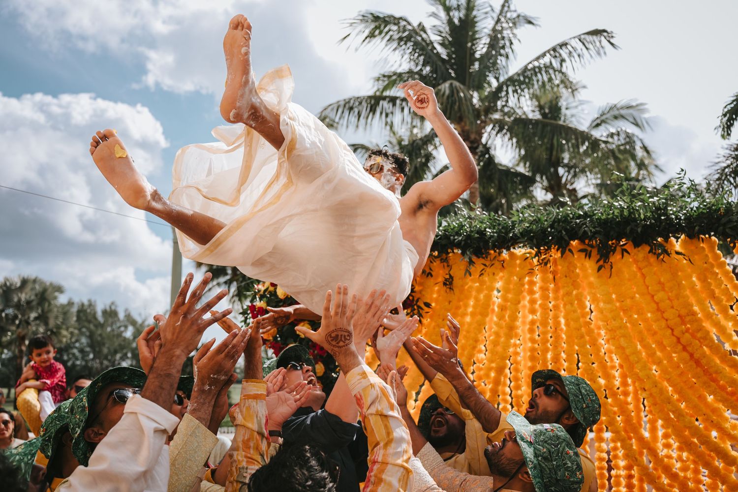 Wedding guests toss and celebrate at an outdoor reception with palm trees and orange string lights in the background.