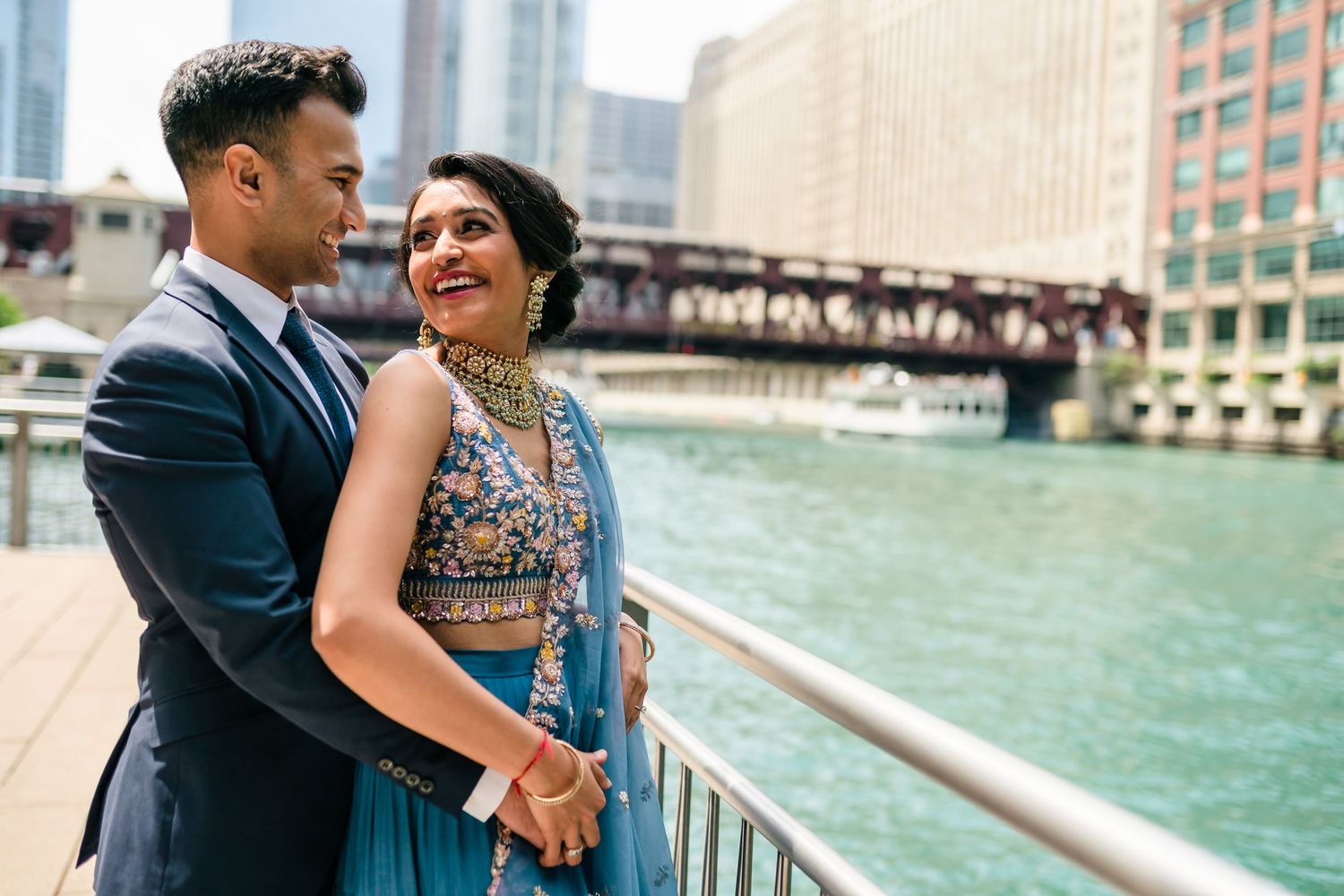 Stylish couple embracing by Chicago River with urban skyline and bridge in background.