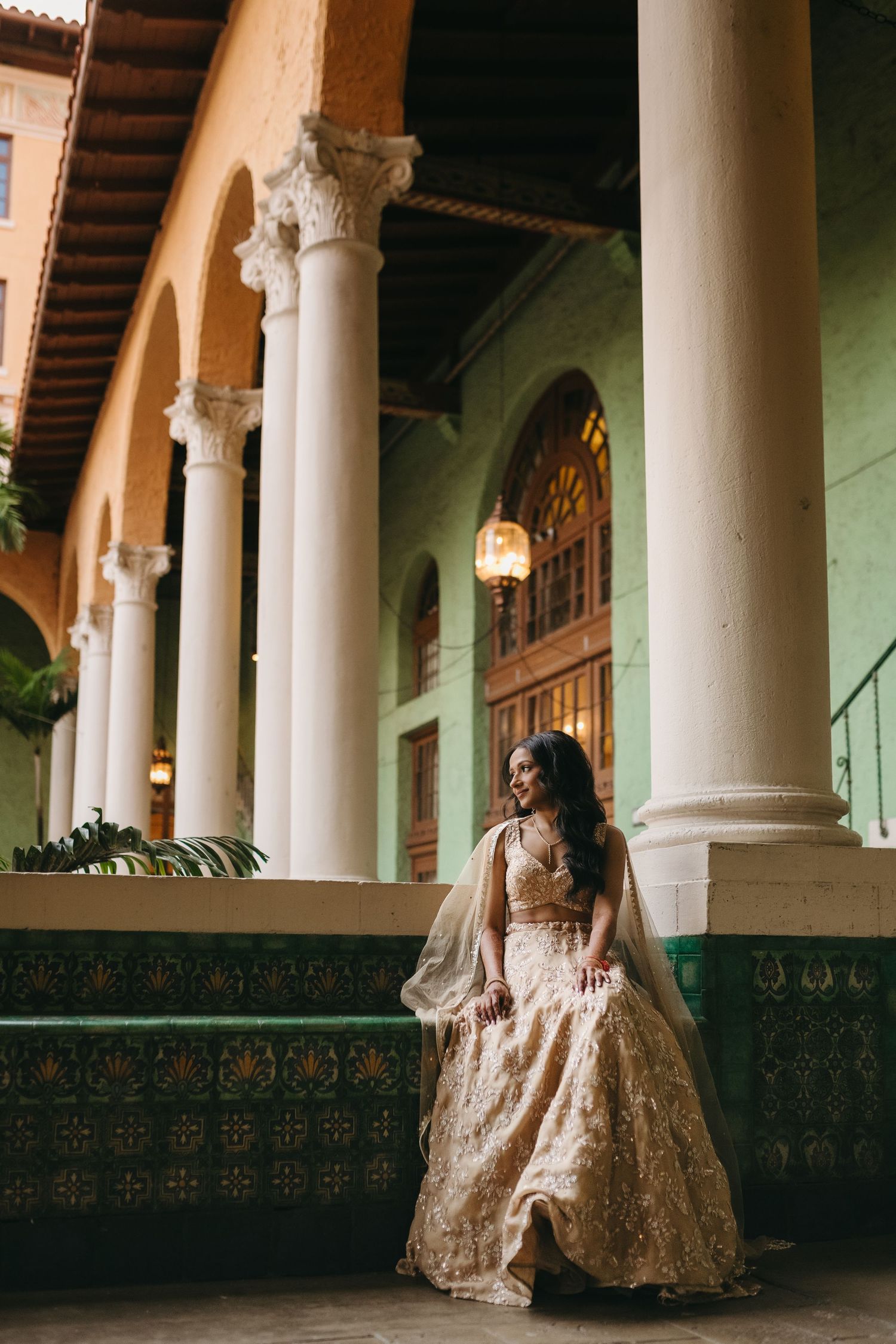 Elegant figure in a champagne lehenga poses against ornate colonial architecture with decorative columns and arches.