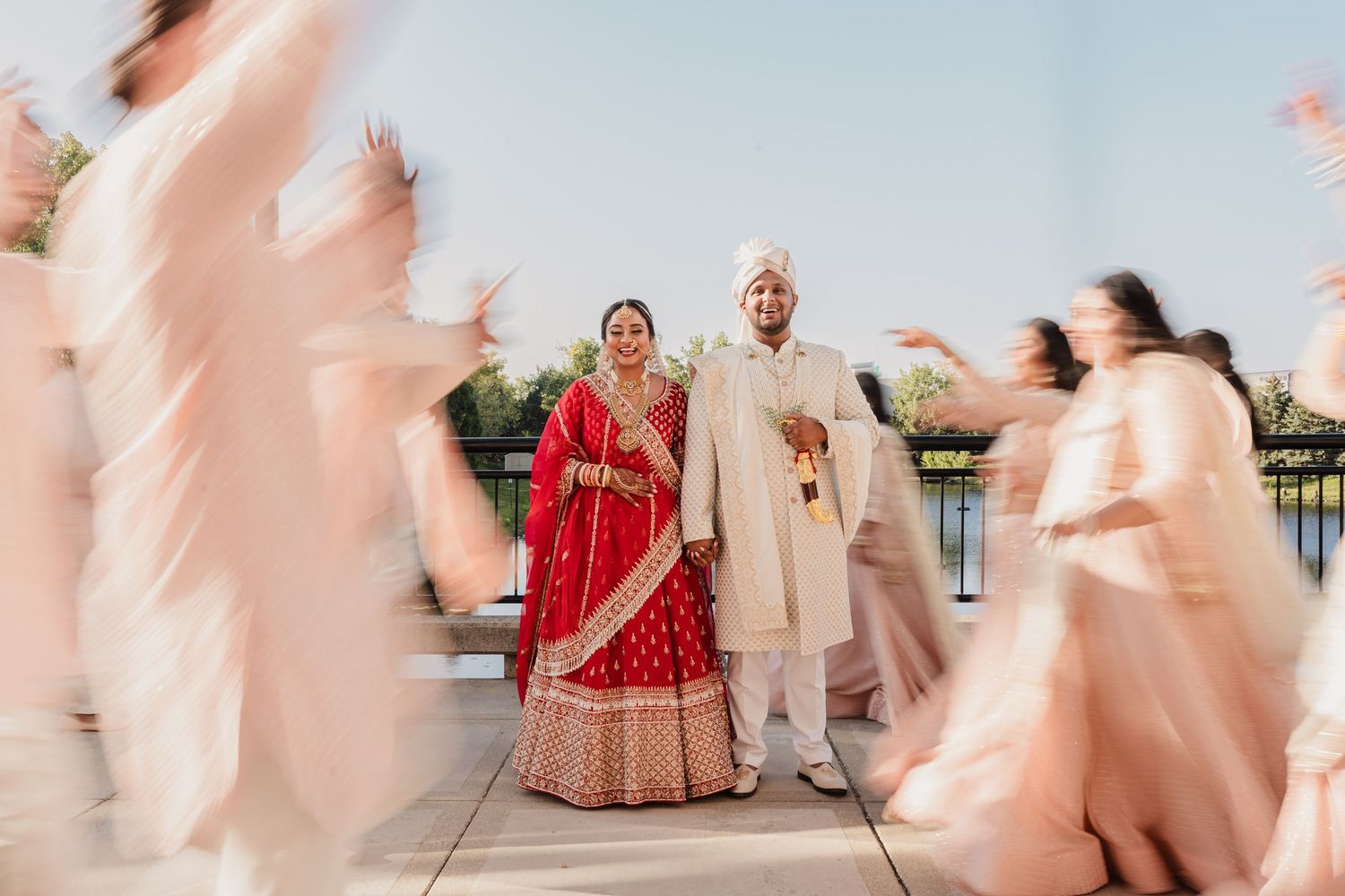 Wedding couple in traditional Indian attire stand amid motion-blurred figures during outdoor celebration.