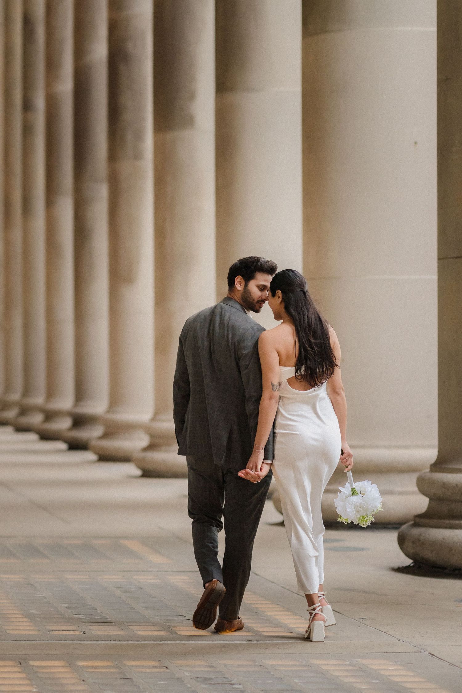 A couple walks together through a colonnade of classical stone pillars.