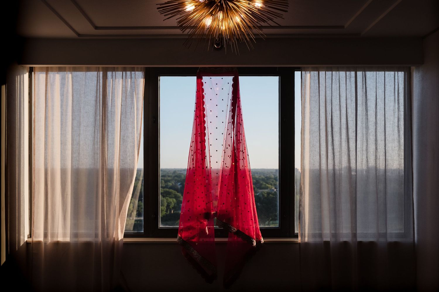 Red fabric draping hangs between white curtains in front of large window with scenic view.