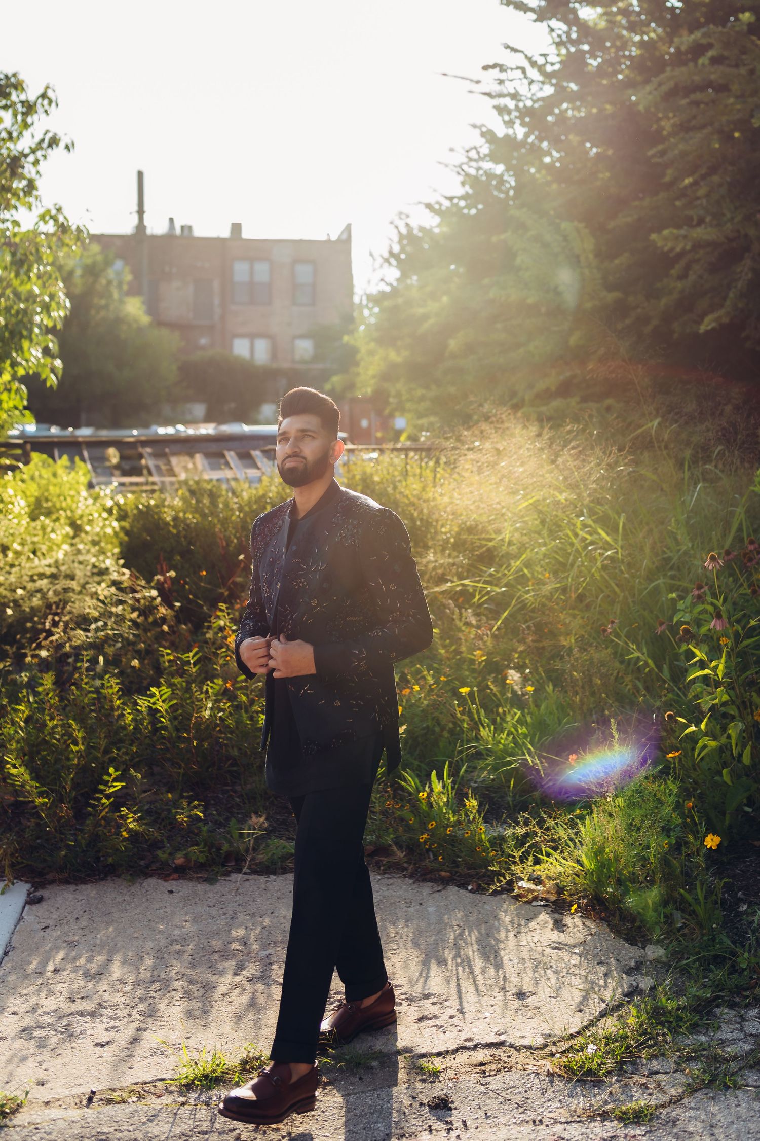 Backlit portrait of man in black attire standing against garden backdrop at sunset.