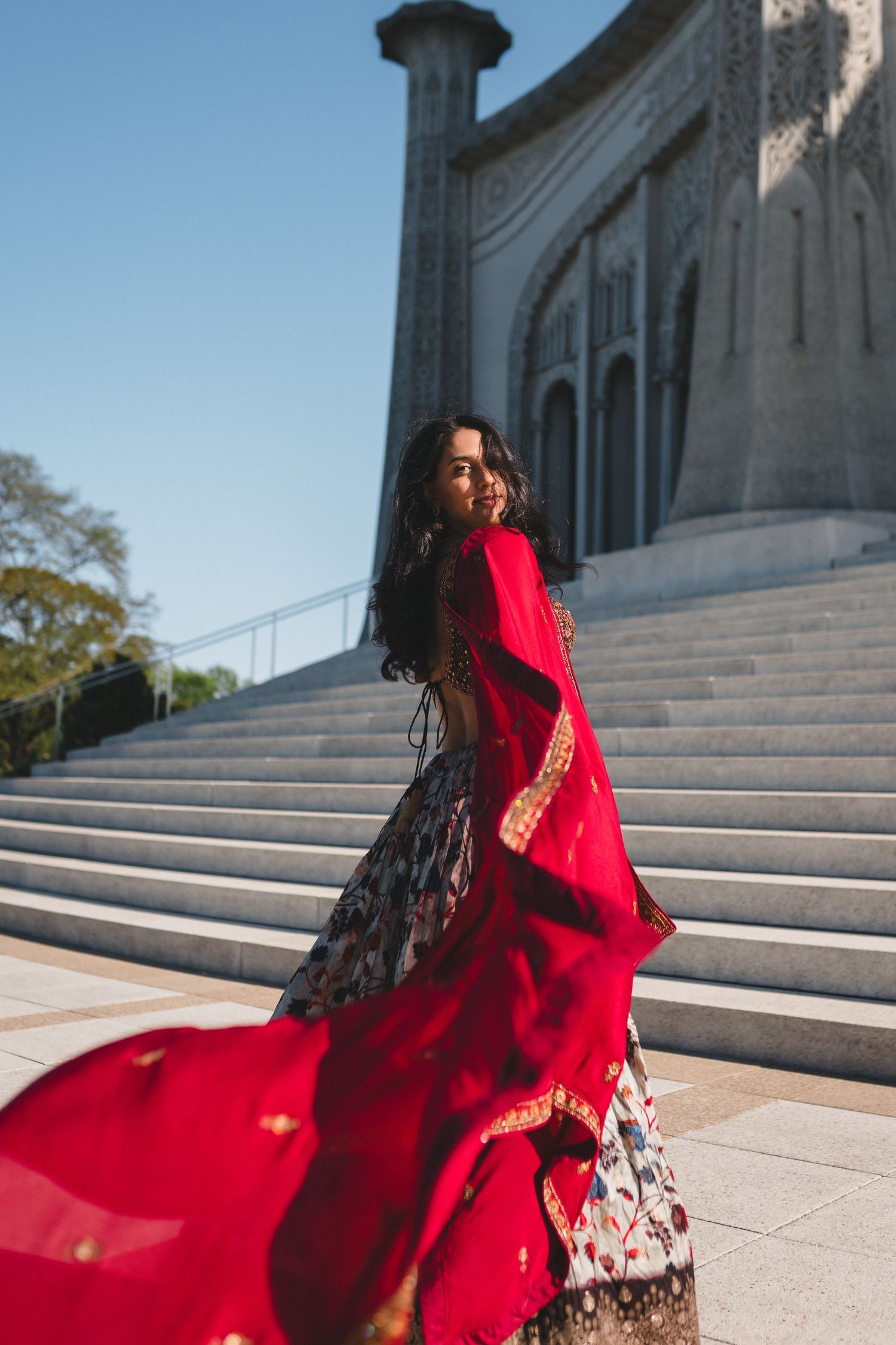 Dramatic red bridal lehenga flows down white marble steps of classical building.