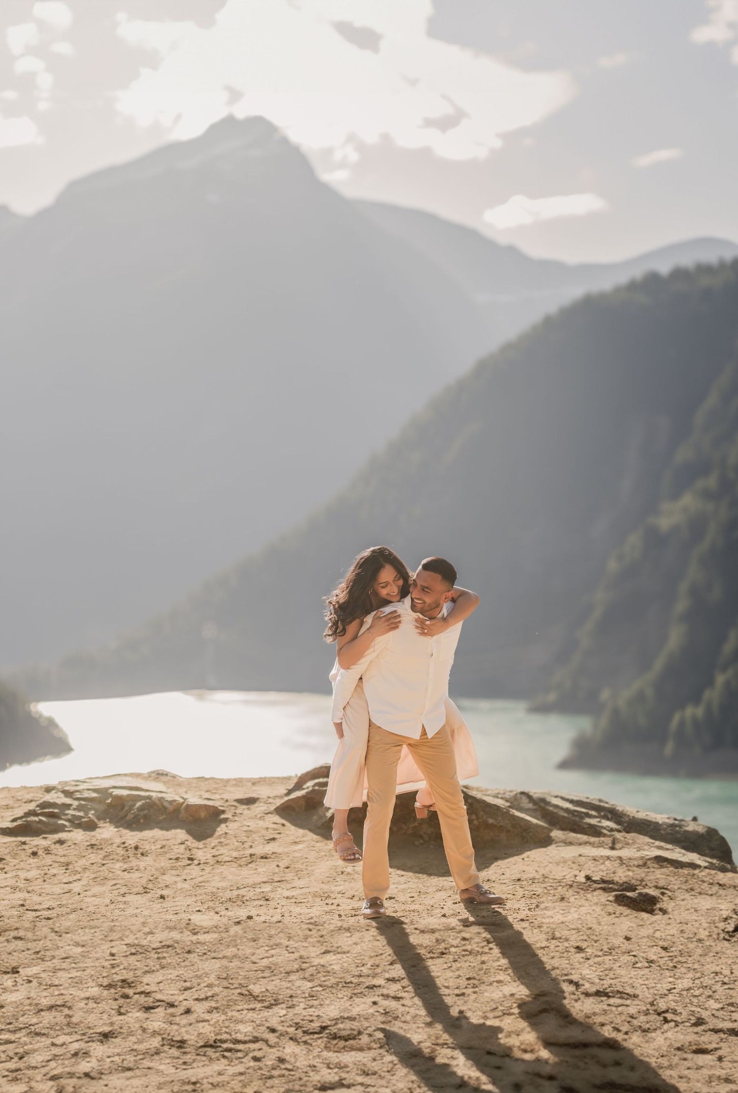 A couple embraces on a mountain cliff overlooking a turquoise lake and misty mountains during sunset.
