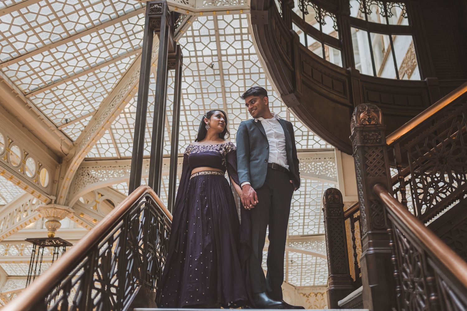 A couple poses on an ornate vintage staircase under a geometric glass ceiling.