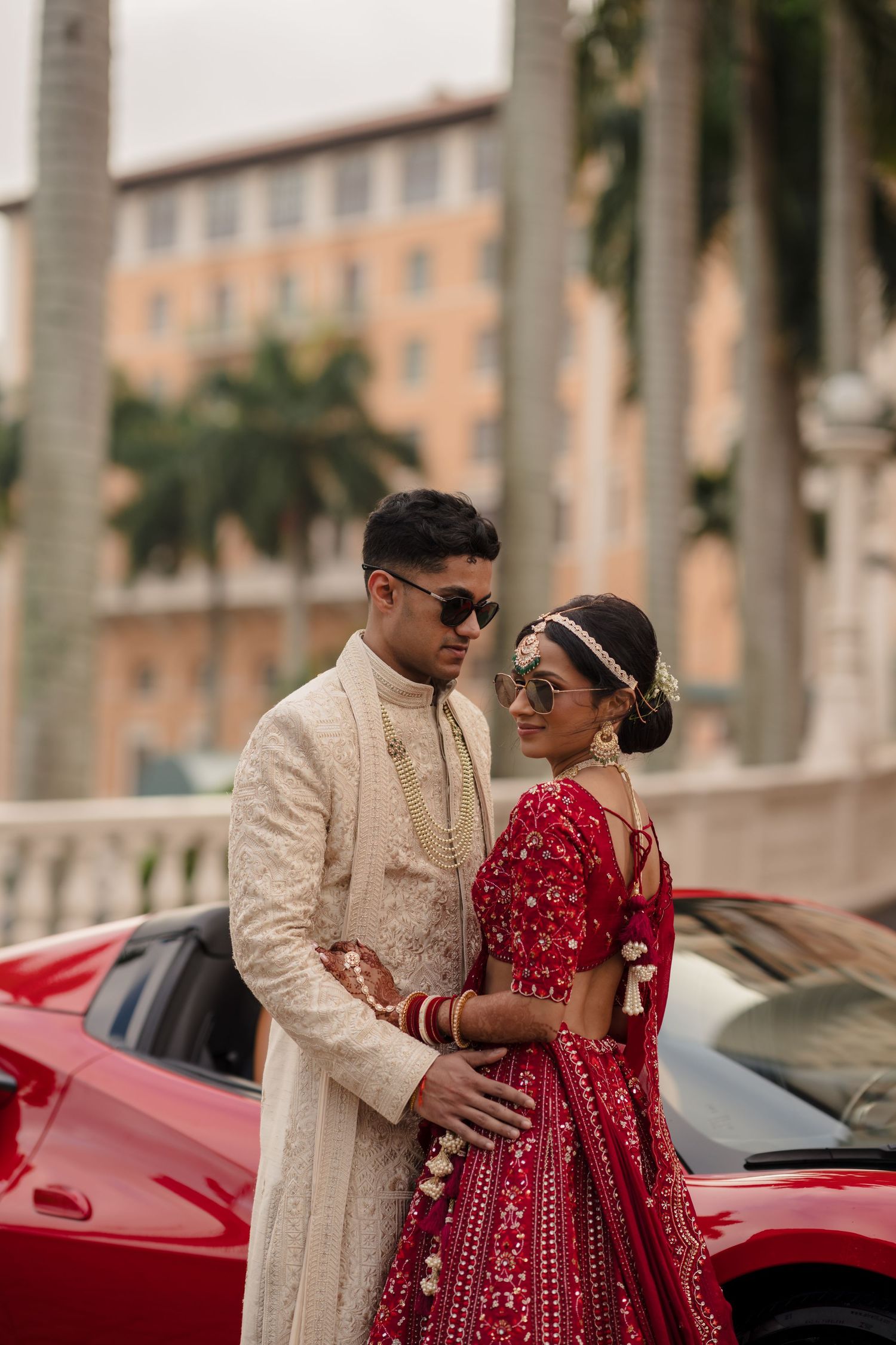 A couple in traditional Indian wedding attire pose together in front of a red car and palm trees.