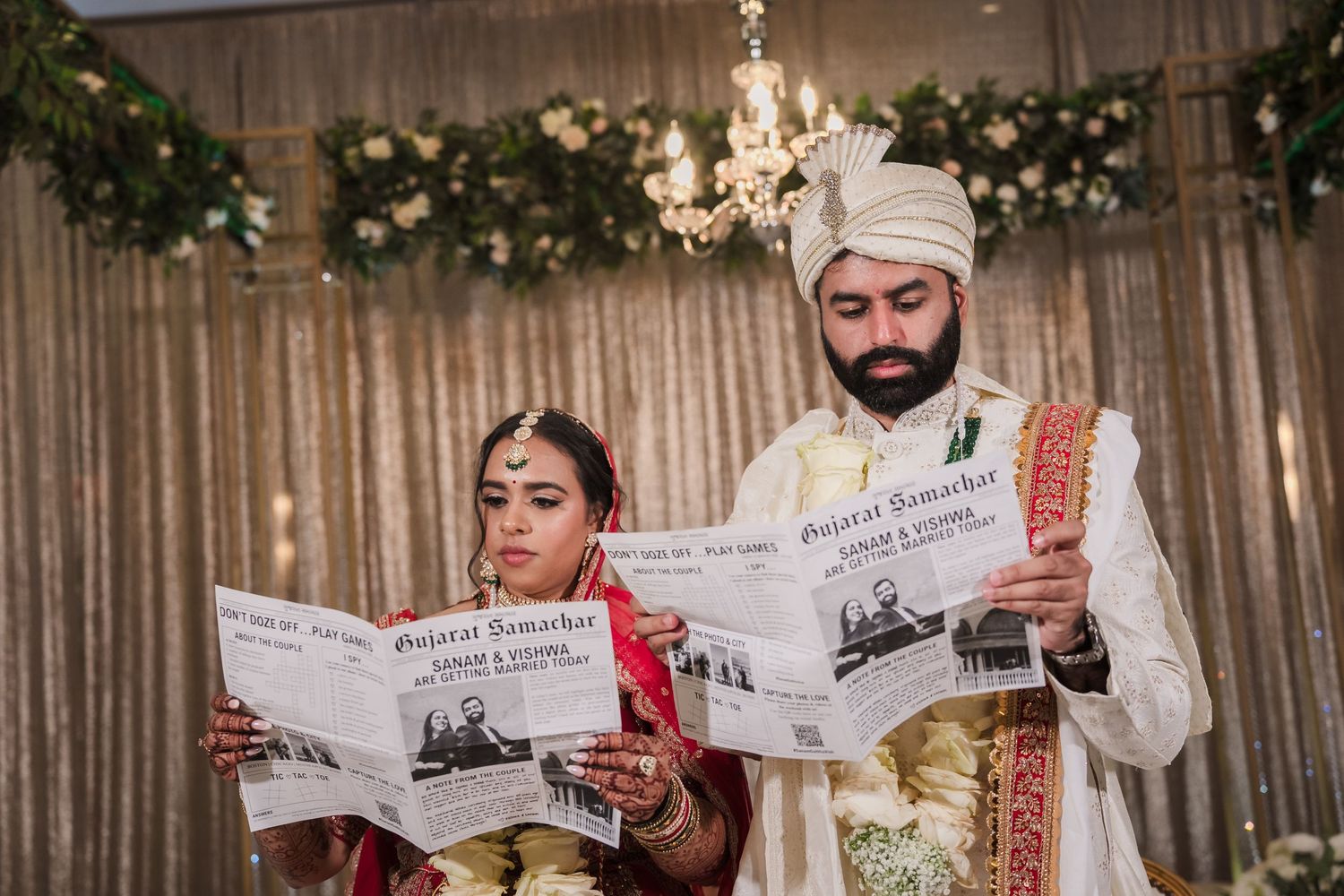 Couple in traditional wedding attire reading newspapers together under chandelier and floral decor.