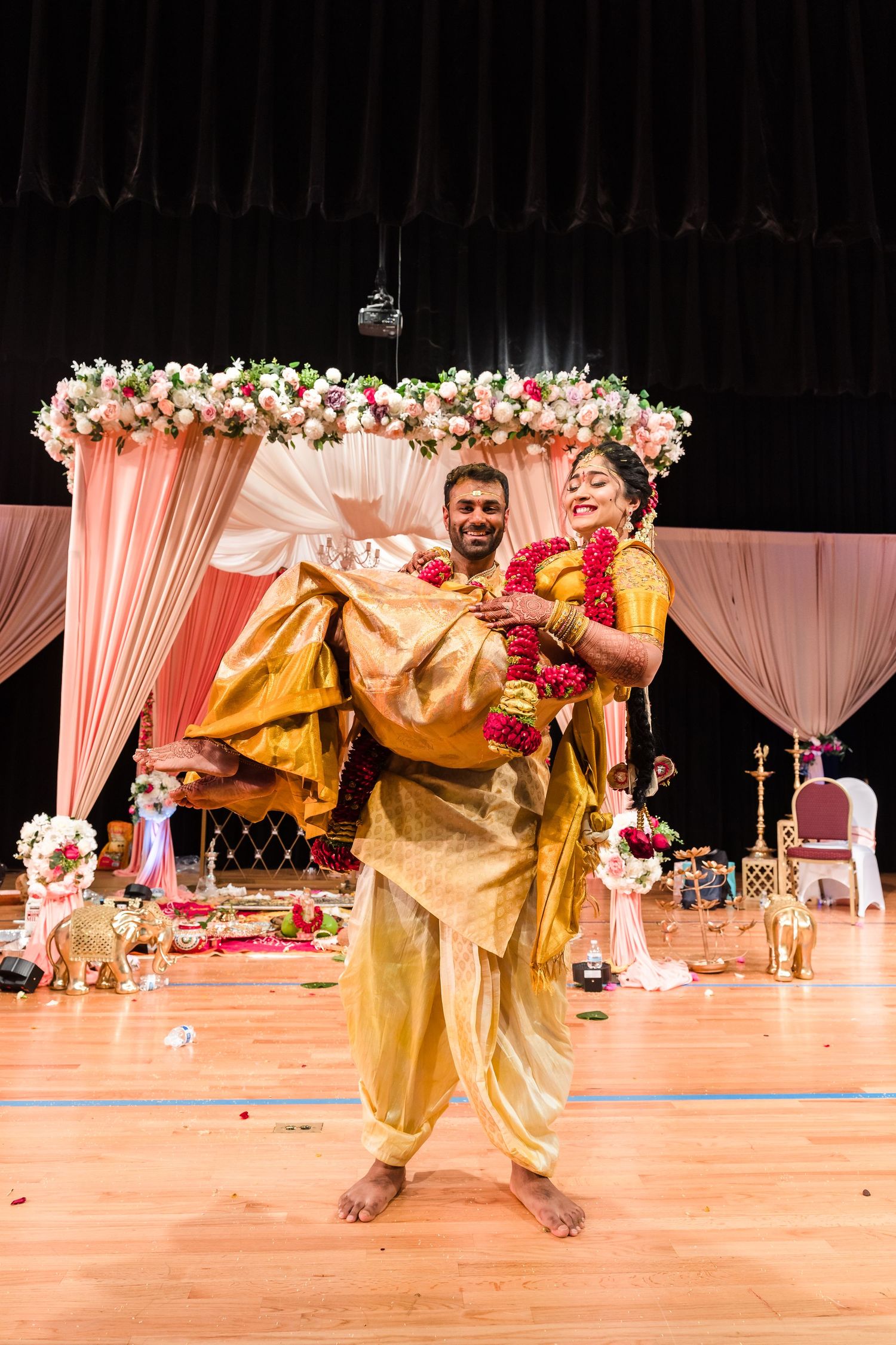 A couple performs a traditional Indian wedding dance in front of a floral mandap decorated in pink and white flowers.
