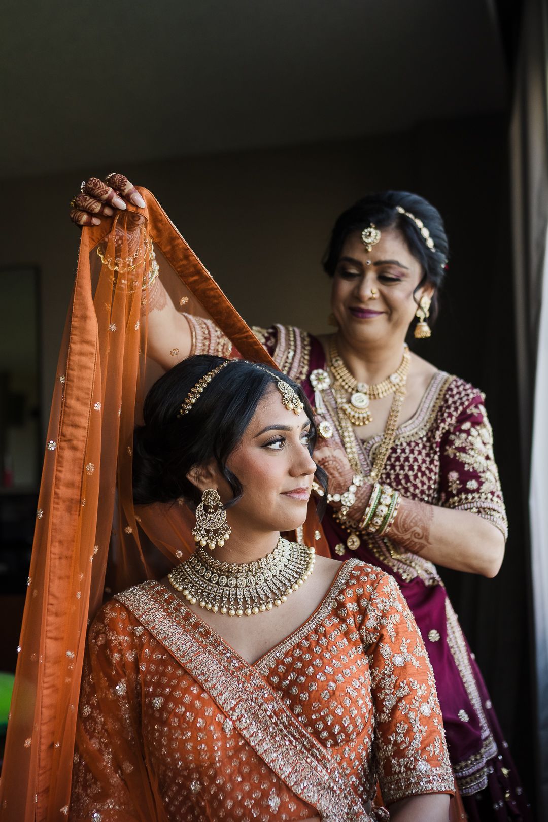 A bride in traditional Indian wedding attire with embellished orange lehenga and jewelry has her dupatta adjusted.