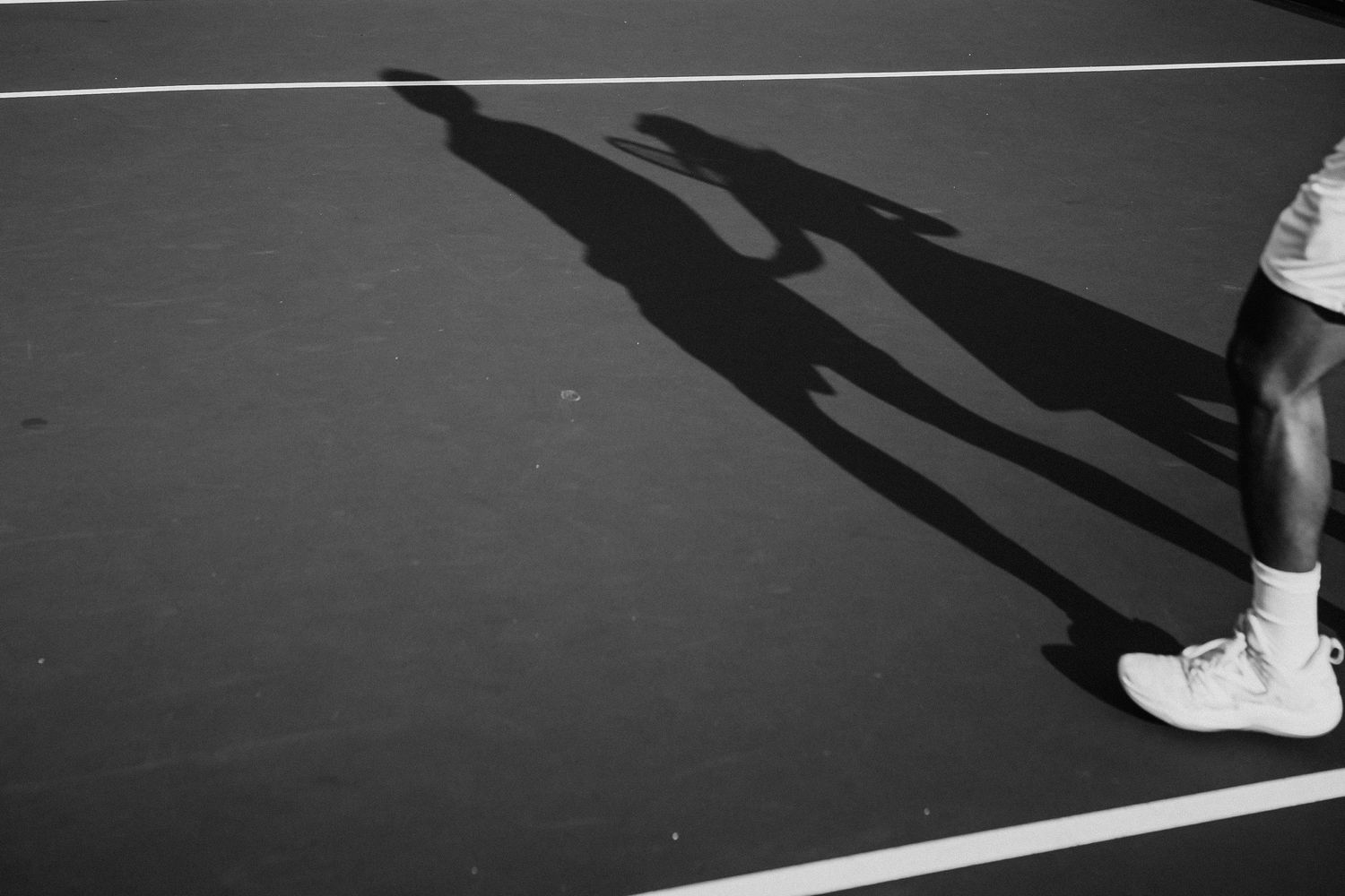 Long shadows cast on a tennis court show players standing near the baseline against a dramatic black and white background.