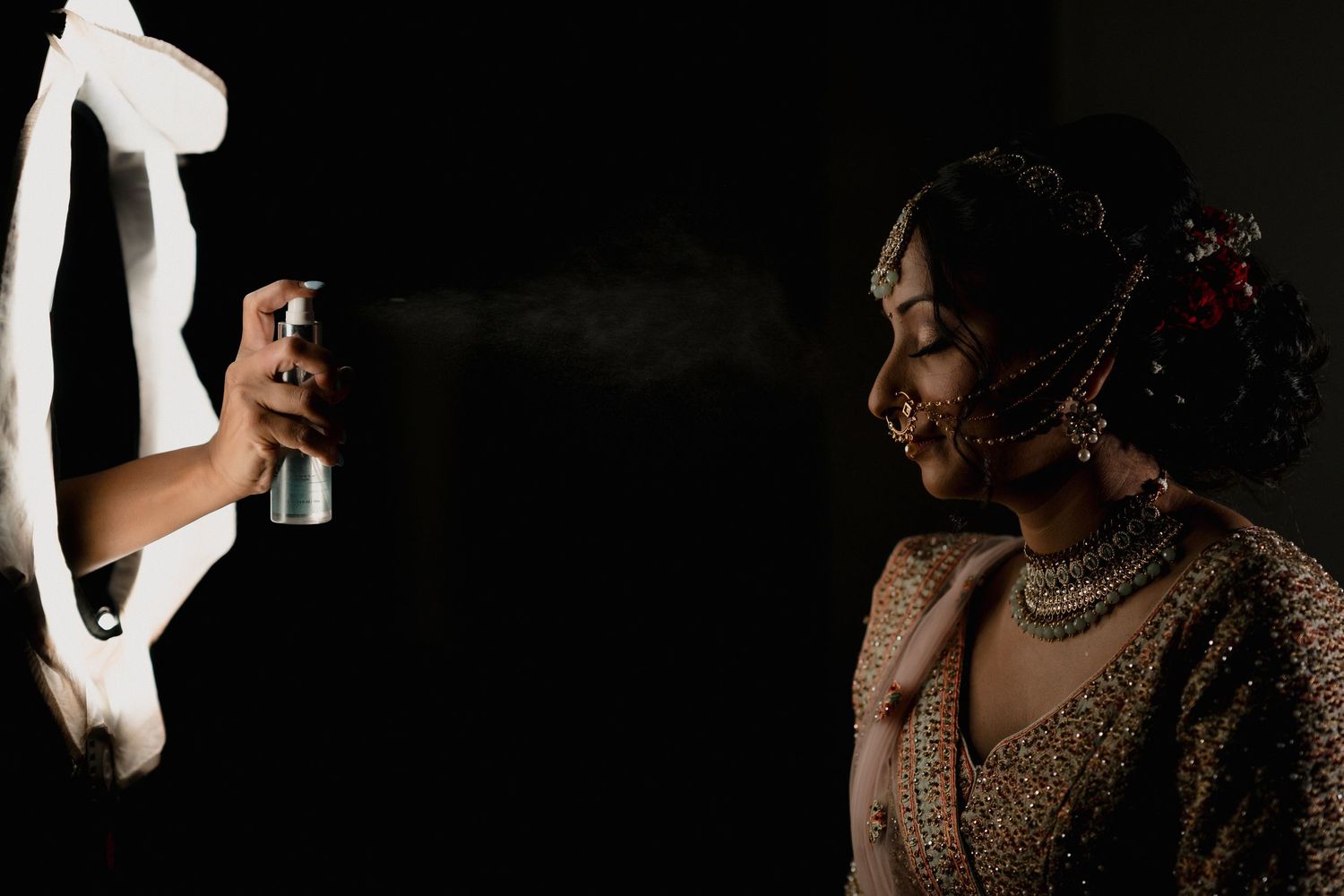 A person in South Asian bridal attire receives a finishing touch of hairspray in a dramatic side-lit photography setup.