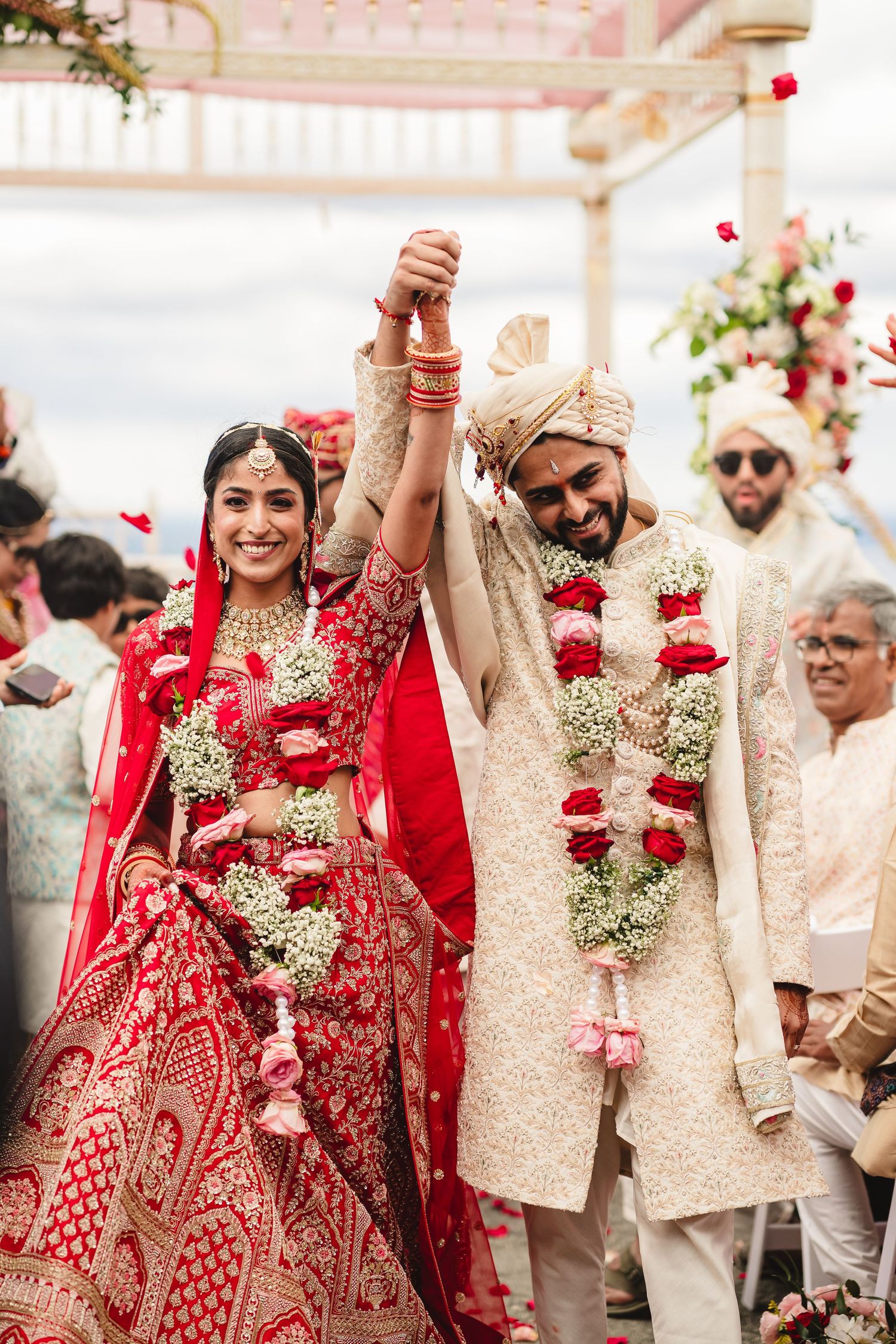 A couple holding hands during a traditional Indian wedding ceremony wearing ornate red and gold attire with floral garlands.