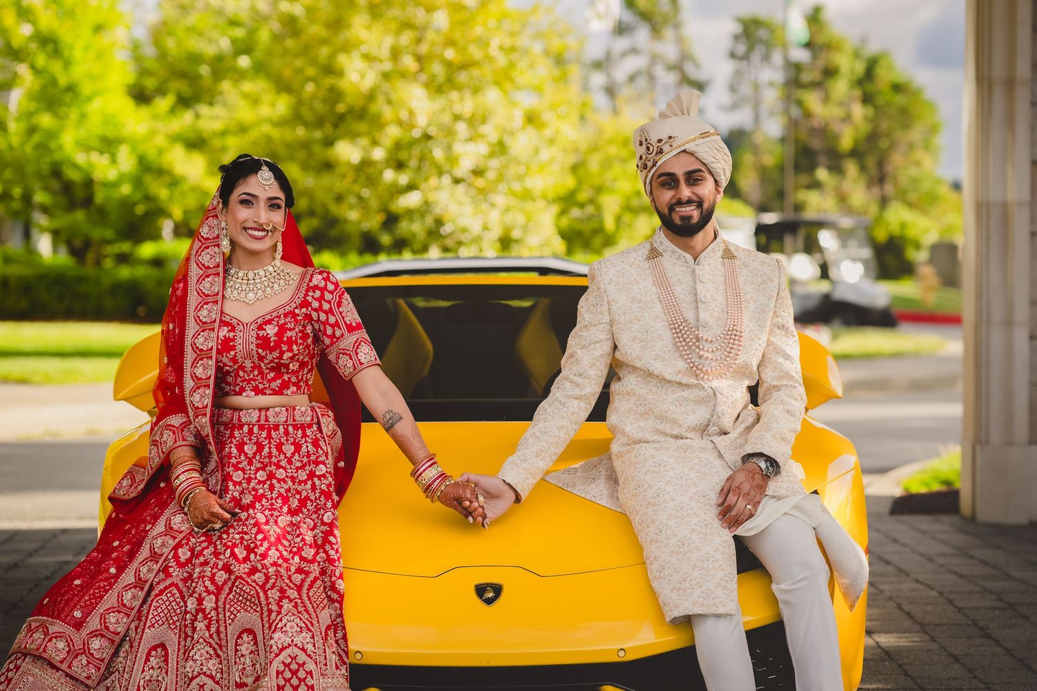 A couple in traditional Indian wedding attire pose together in front of a bright yellow sports car on a sunny day.