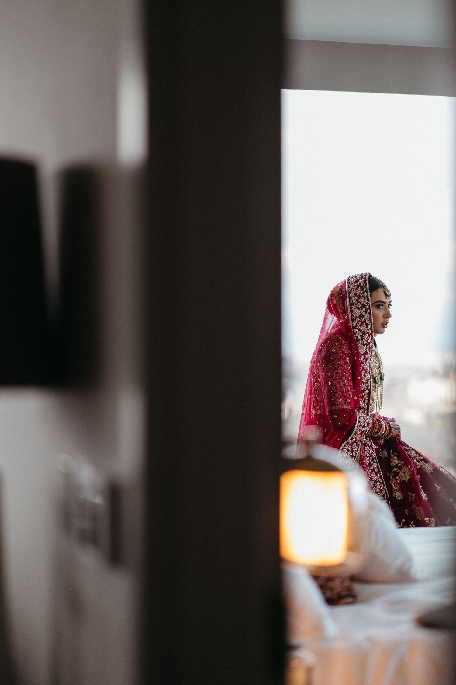 Silhouette of a bride in traditional red attire sitting by a window with soft lighting.
