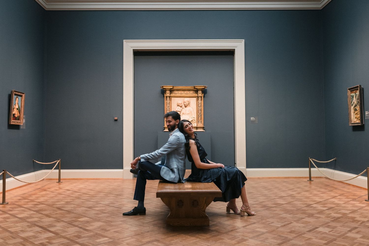 Two visitors sit on a bench in an art museum gallery with blue walls and white doorways.