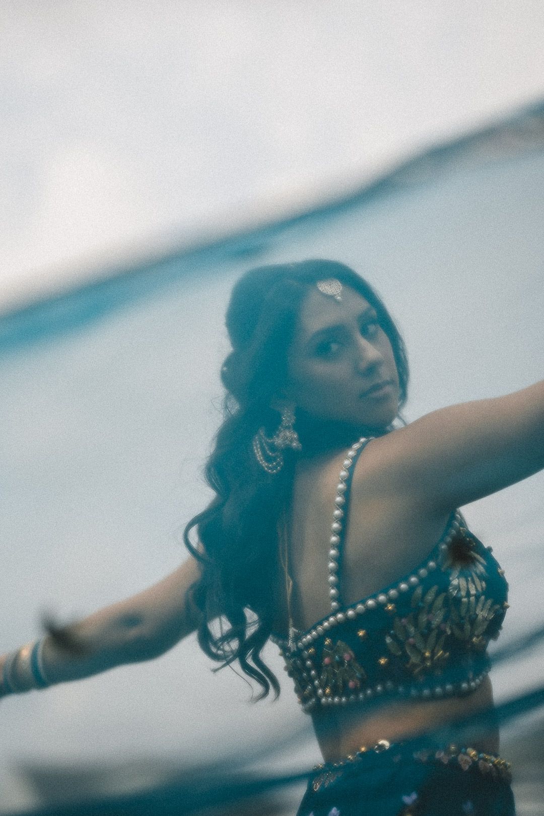 A dancer in an embellished crop top performs with graceful outstretched arms in a dreamy blue-tinted setting.