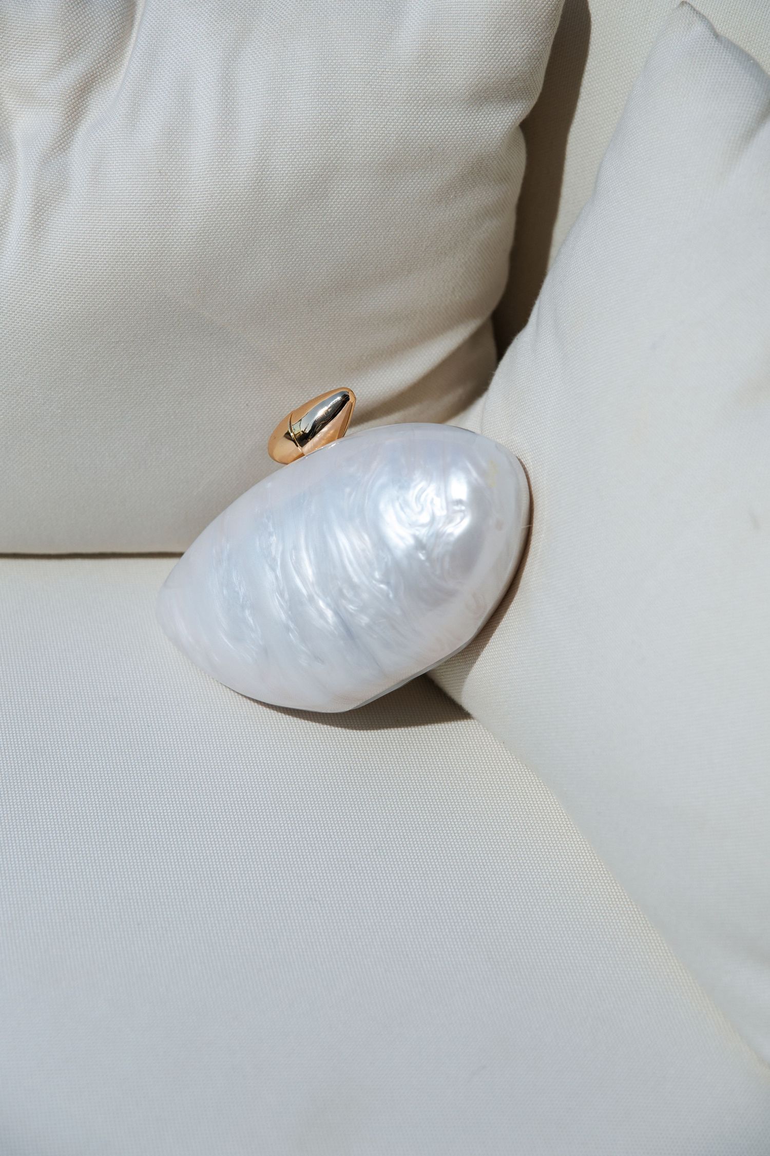 A pearlescent white shell-shaped clutch handbag with gold trim rests on a light-colored couch or cushion.
