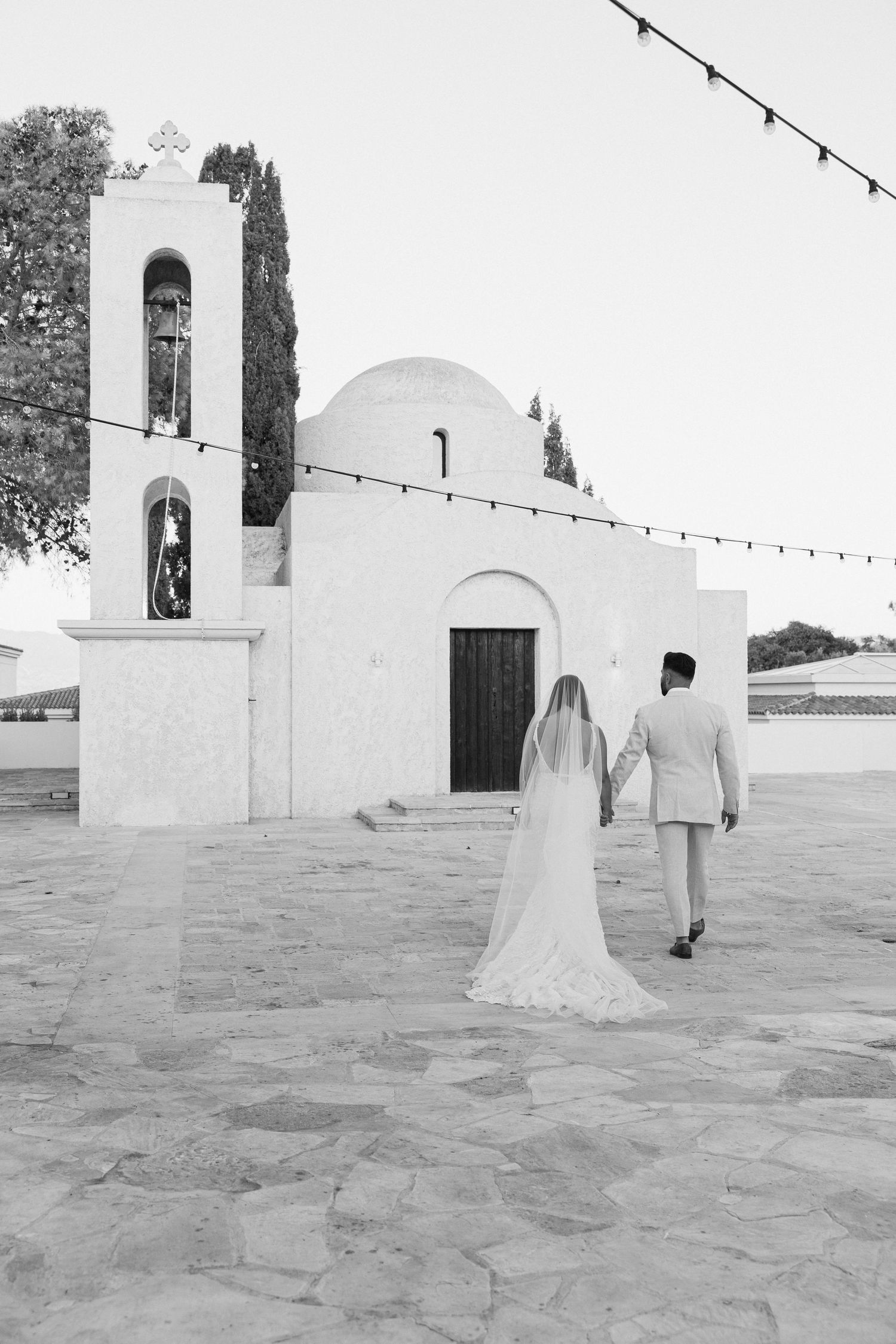 Bride and groom walk hand in hand toward a white Greek chapel with bell tower in black and white photo.