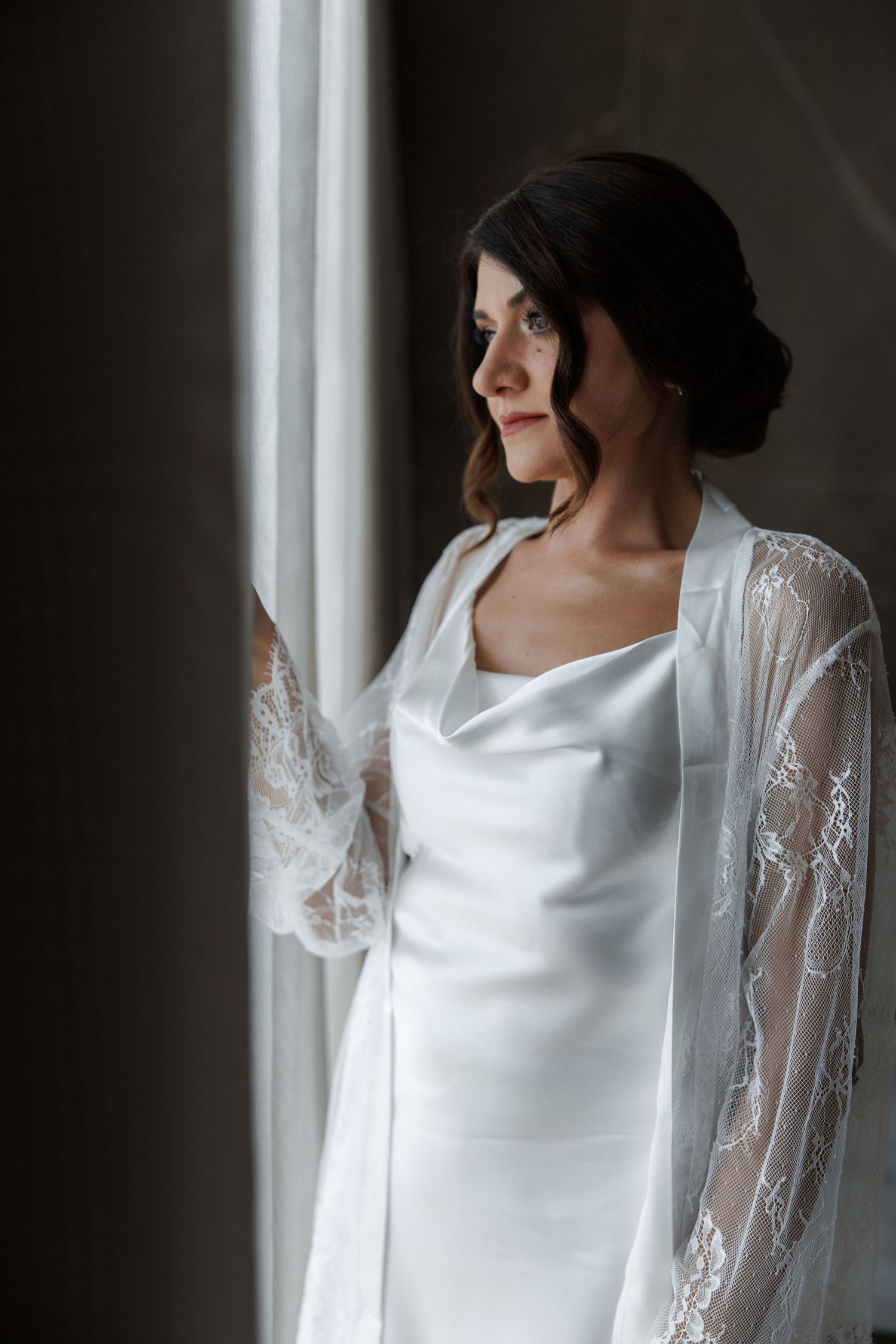 Bride in white satin gown and lace robe gazes out window in soft natural light.