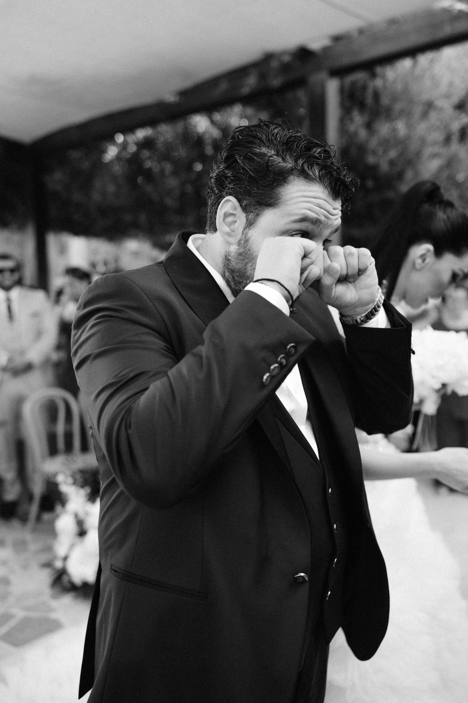 Emotional groom wiping tears at outdoor wedding ceremony, wearing dark suit, black and white photo.