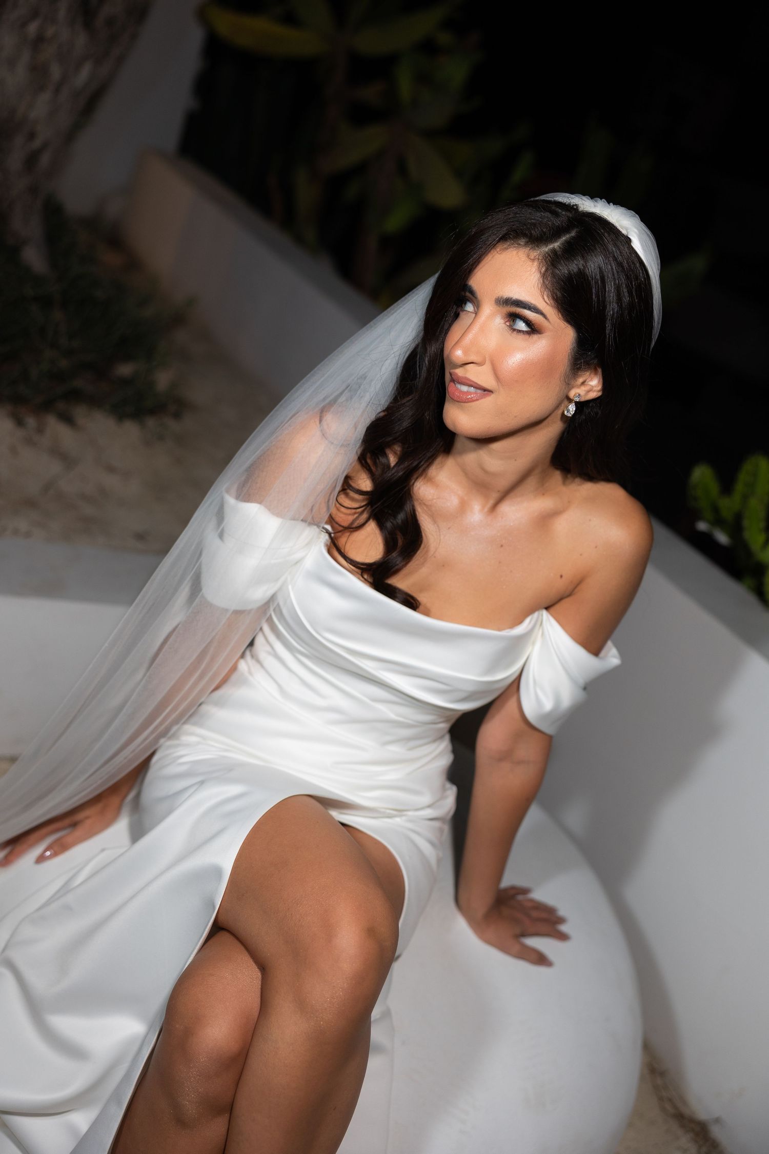 Elegant bride in off-shoulder white gown with veil and headband sits outdoors at night wedding.