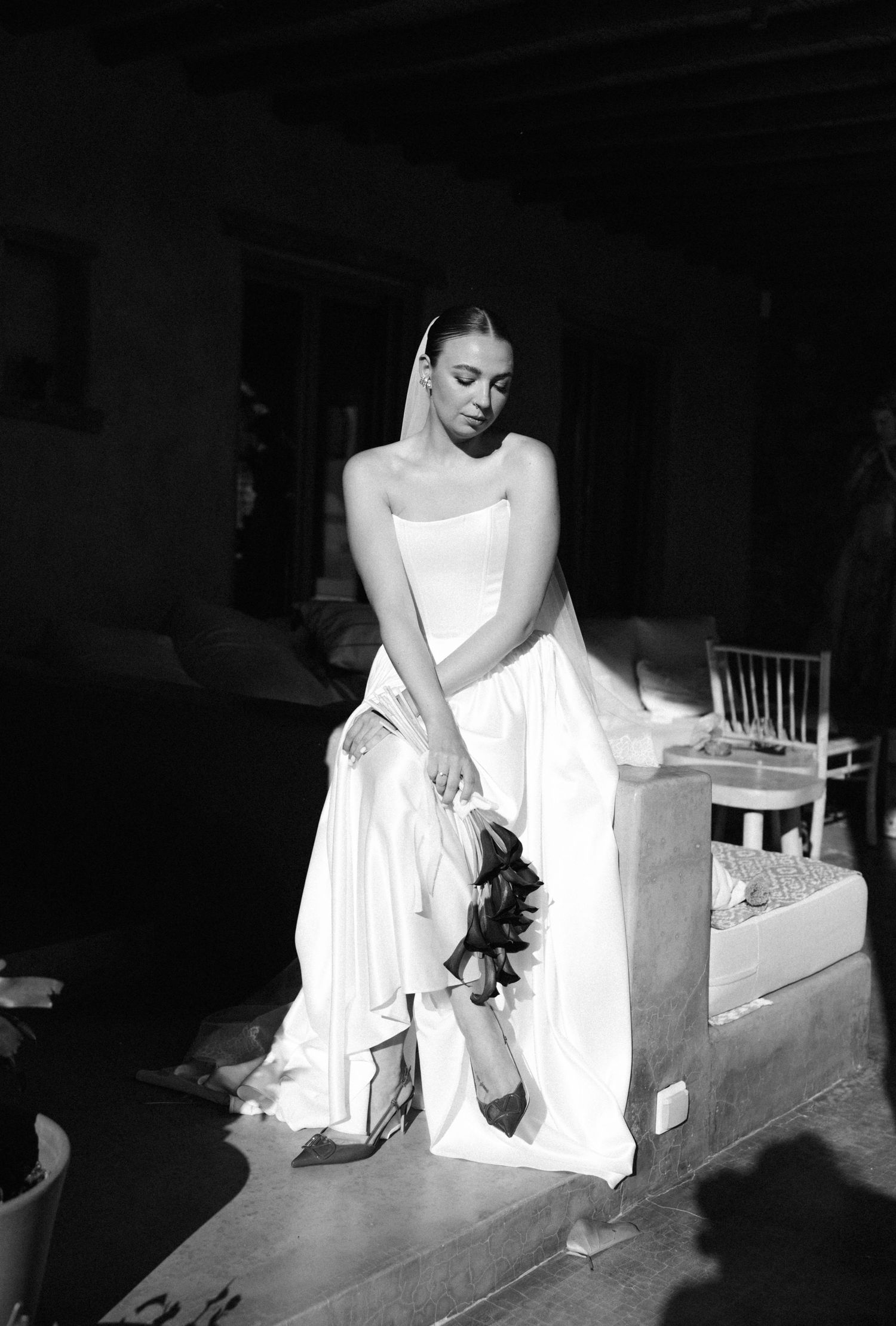 Black and white photo of a bride in a strapless gown adjusting her heels while holding a bouquet.