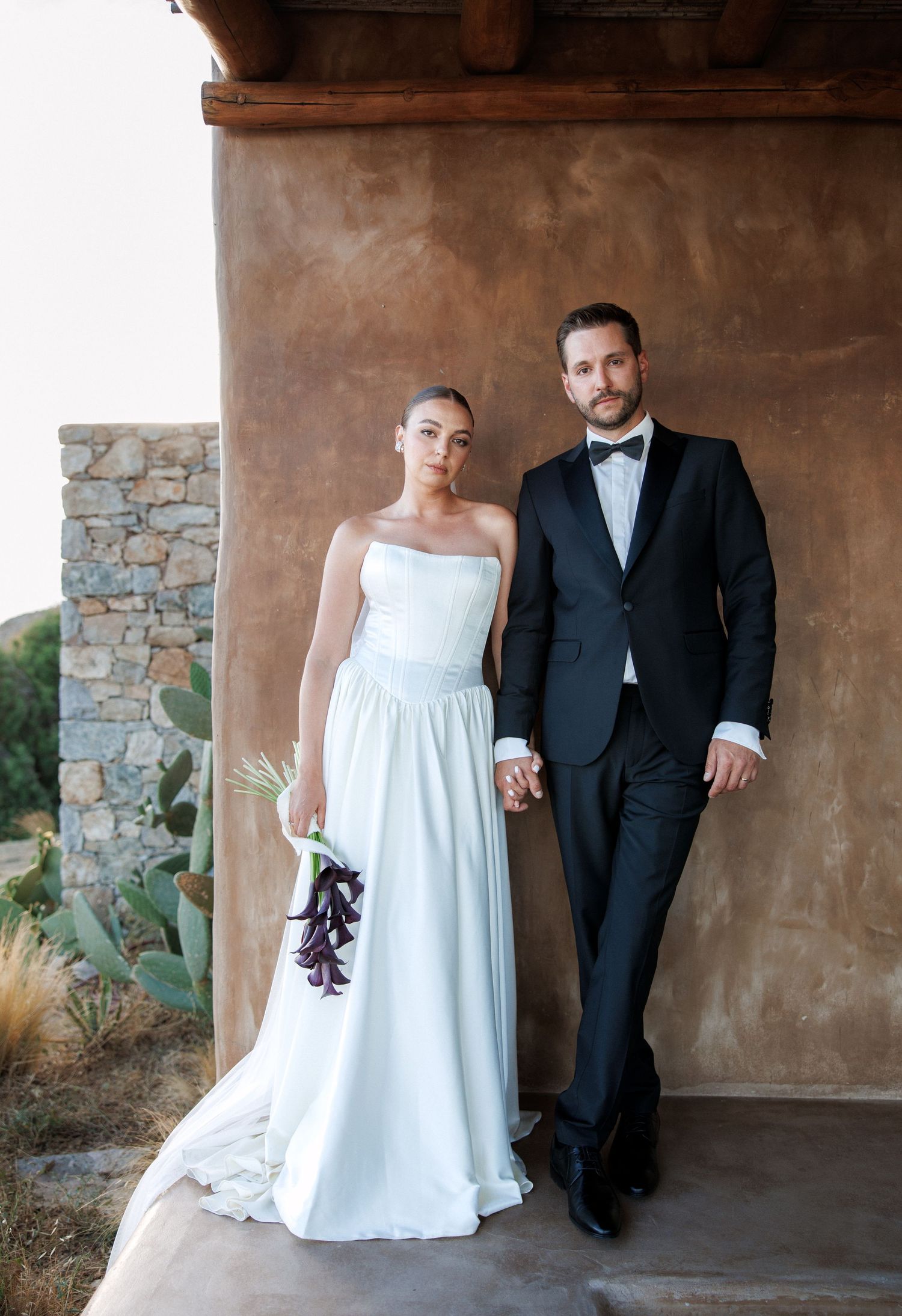 Bride in strapless white gown and groom in black tuxedo pose together holding a dark floral bouquet.