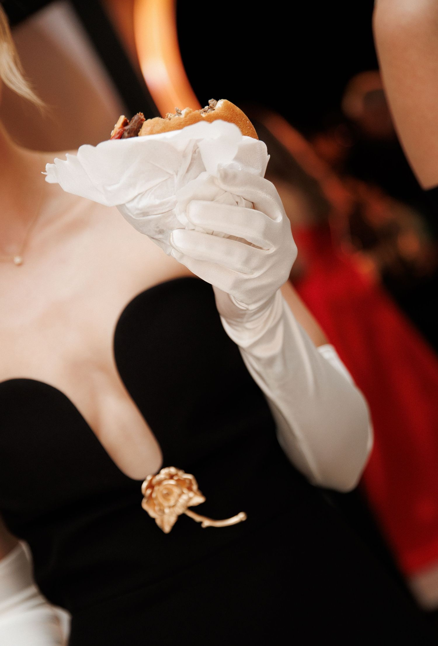 Elegant woman in black strapless gown with white satin gloves holding a burger at a formal event.