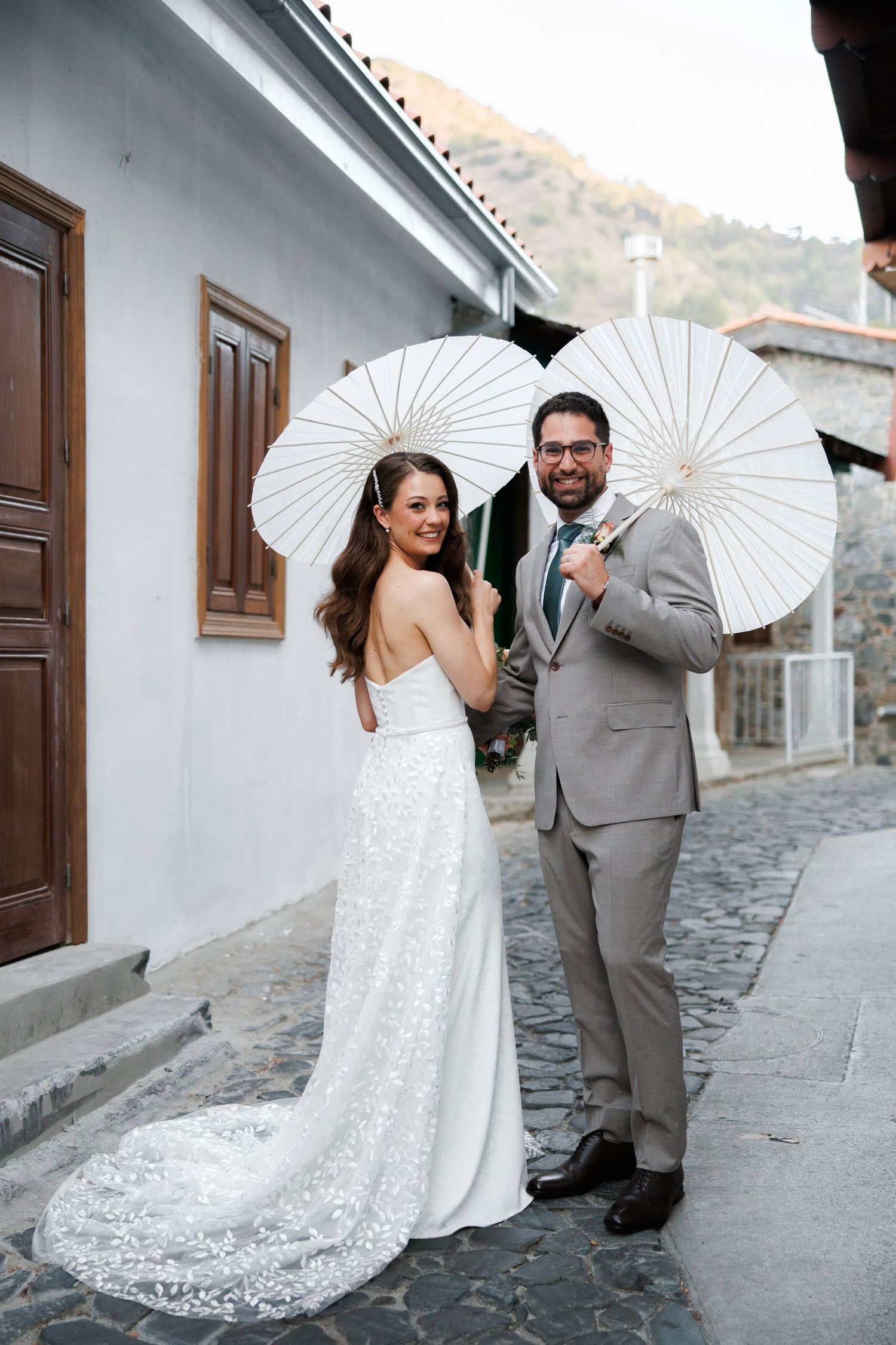 Newlyweds posing with white parasols against white wall in rustic setting.