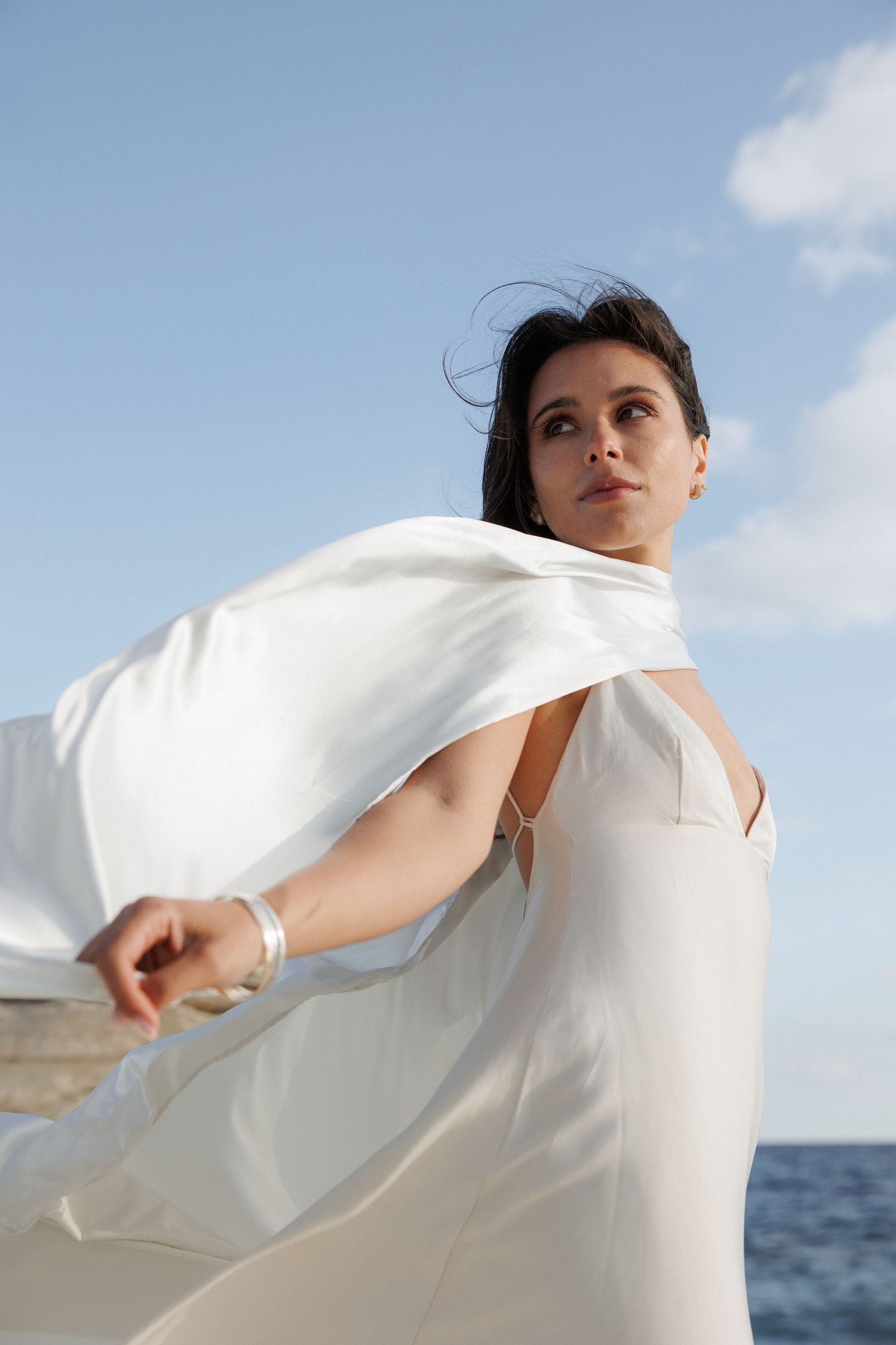 Figure in flowing white dress moves gracefully against ocean backdrop with blue sky and clouds above.