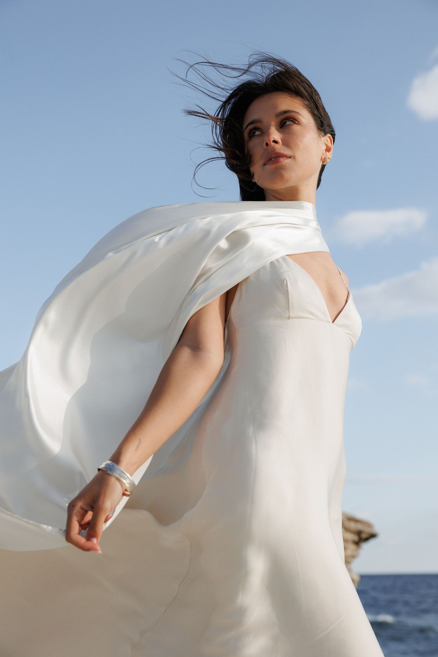 Figure in flowing white dress with windswept hair against blue sky and ocean backdrop.