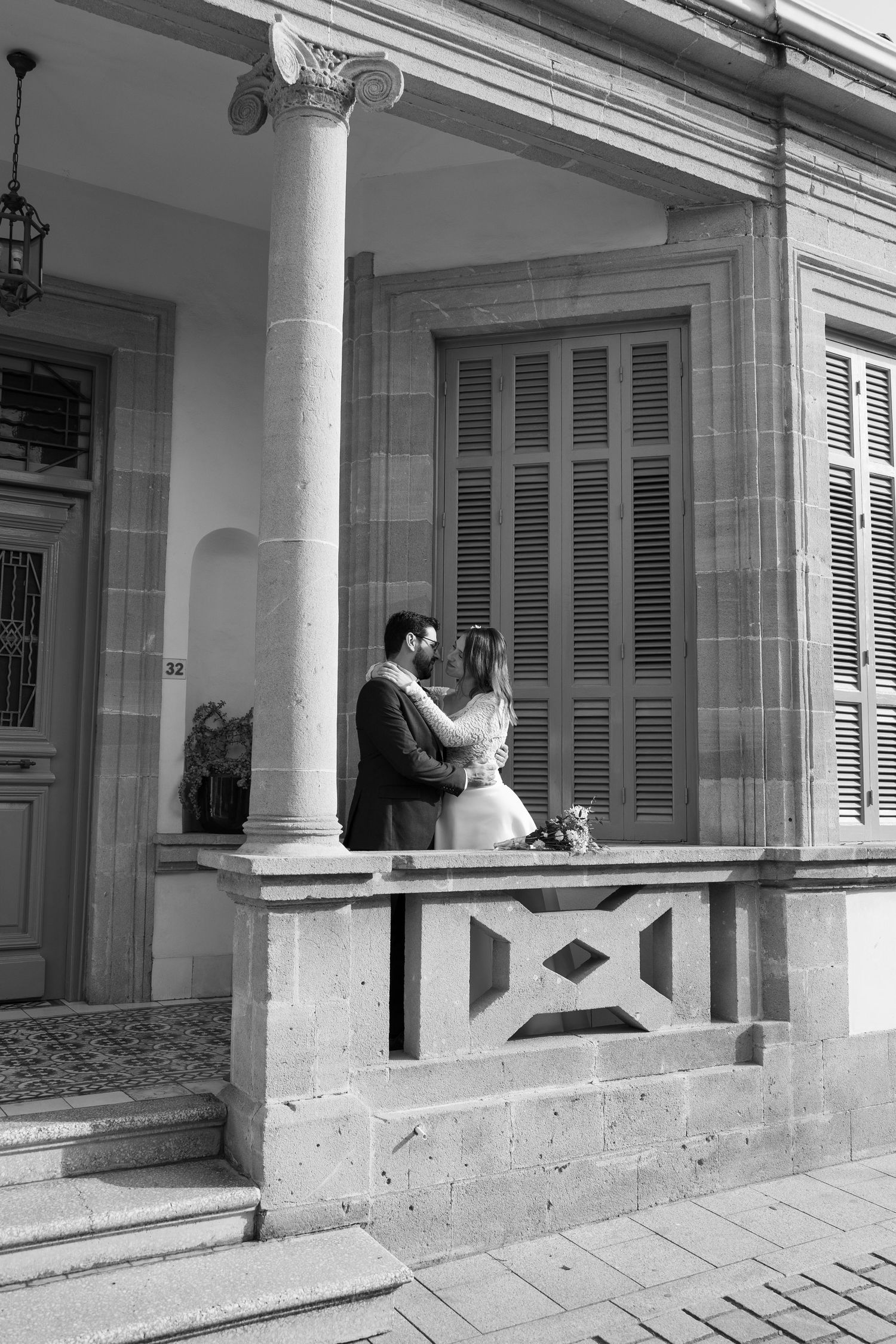 Black and white photo of newlyweds embracing on elegant stone building portico with classical columns.