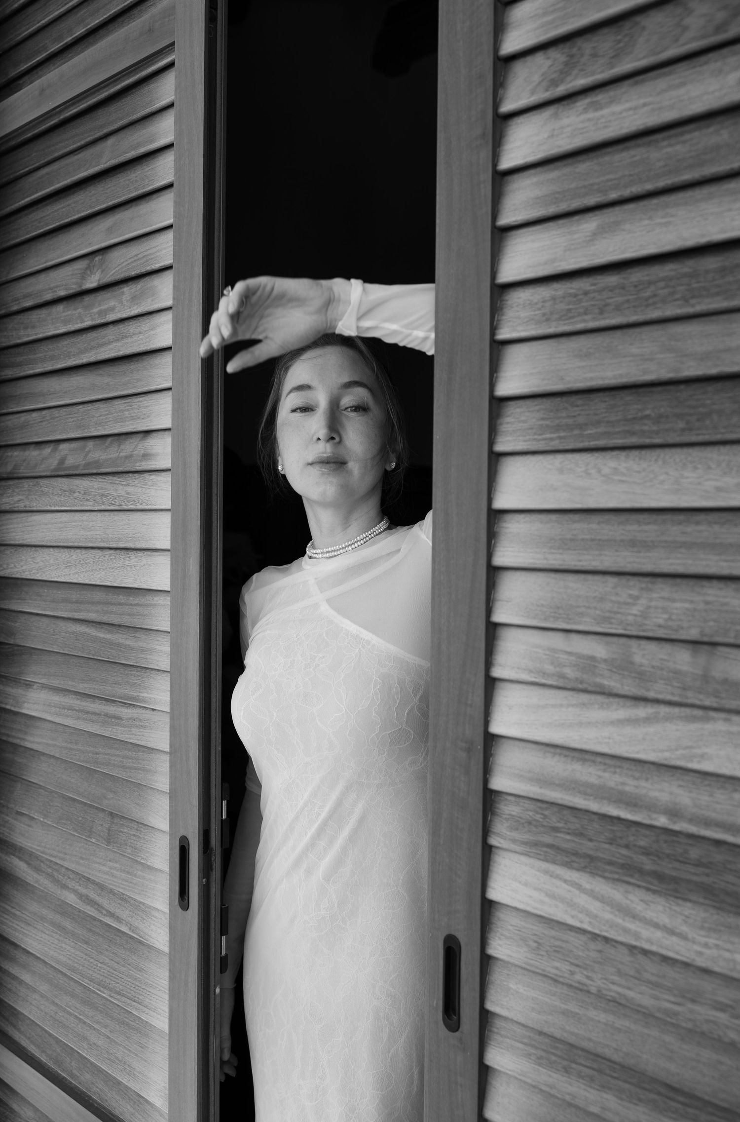 Black and white photo of a woman in a white dress posing in a wooden doorway with her arm raised.