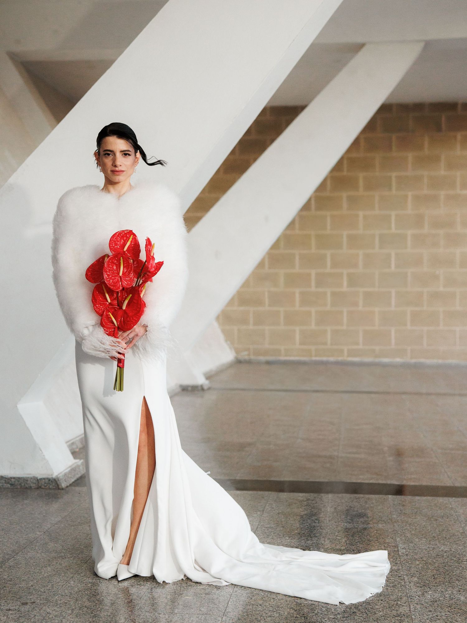 Bride in white gown with fluffy fur wrap holding red anthurium bouquet, standing in modern architectural space.