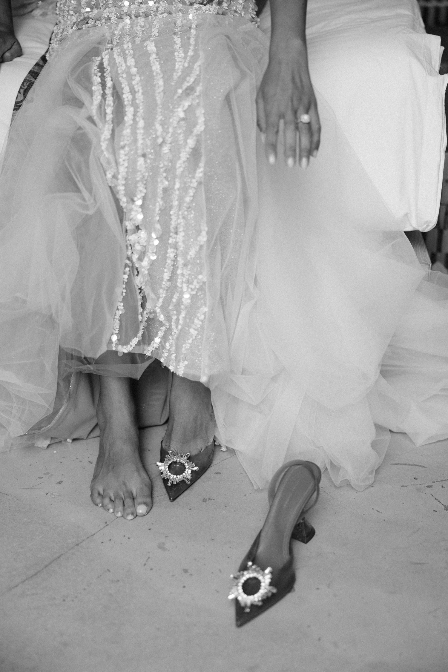 Black and white photo of two women in elegant gowns, one barefoot, one in jeweled heels at a formal event.