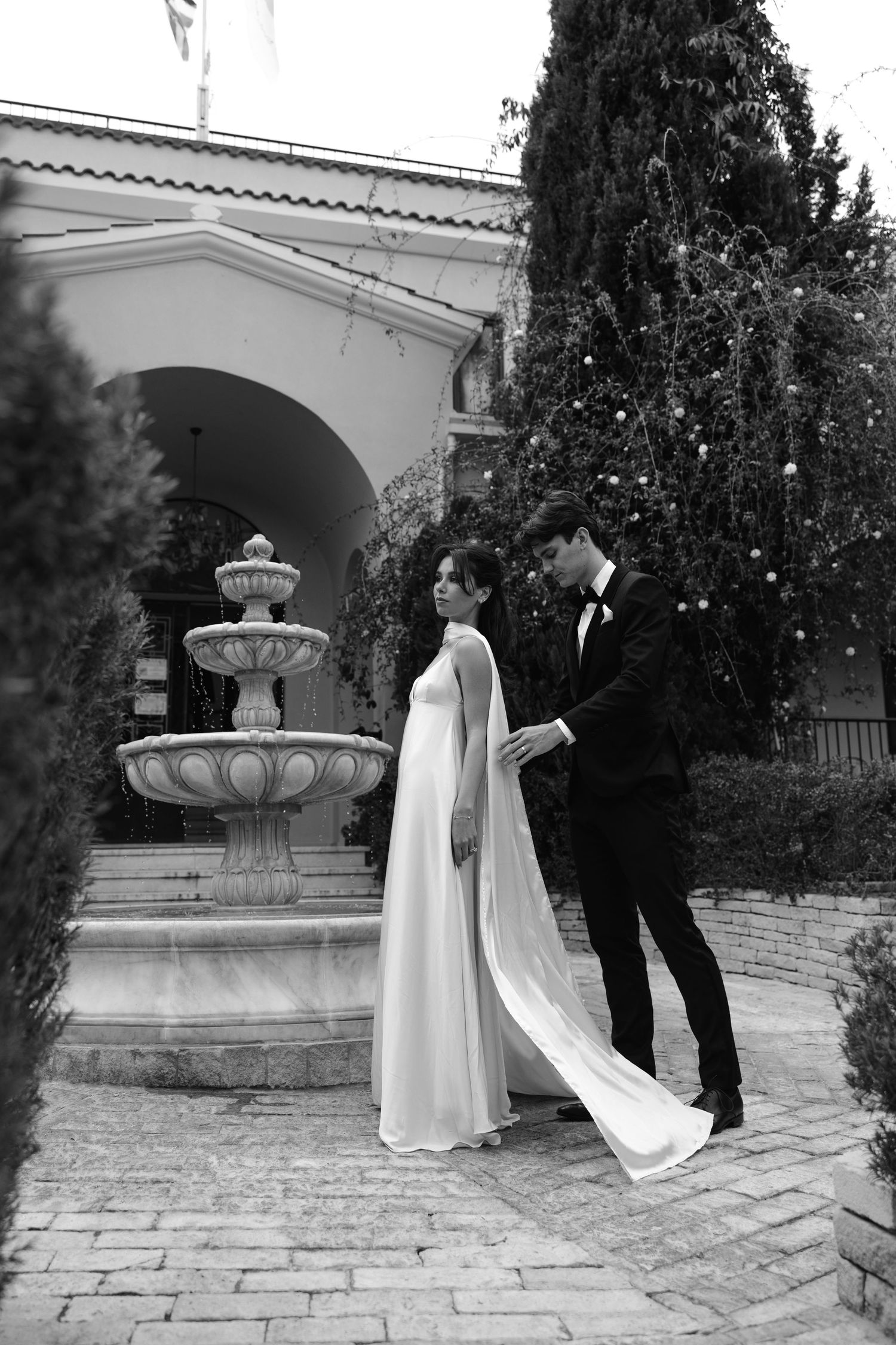Black and white wedding photo with newlyweds standing next to an elegant stone fountain in front of an arched doorway.